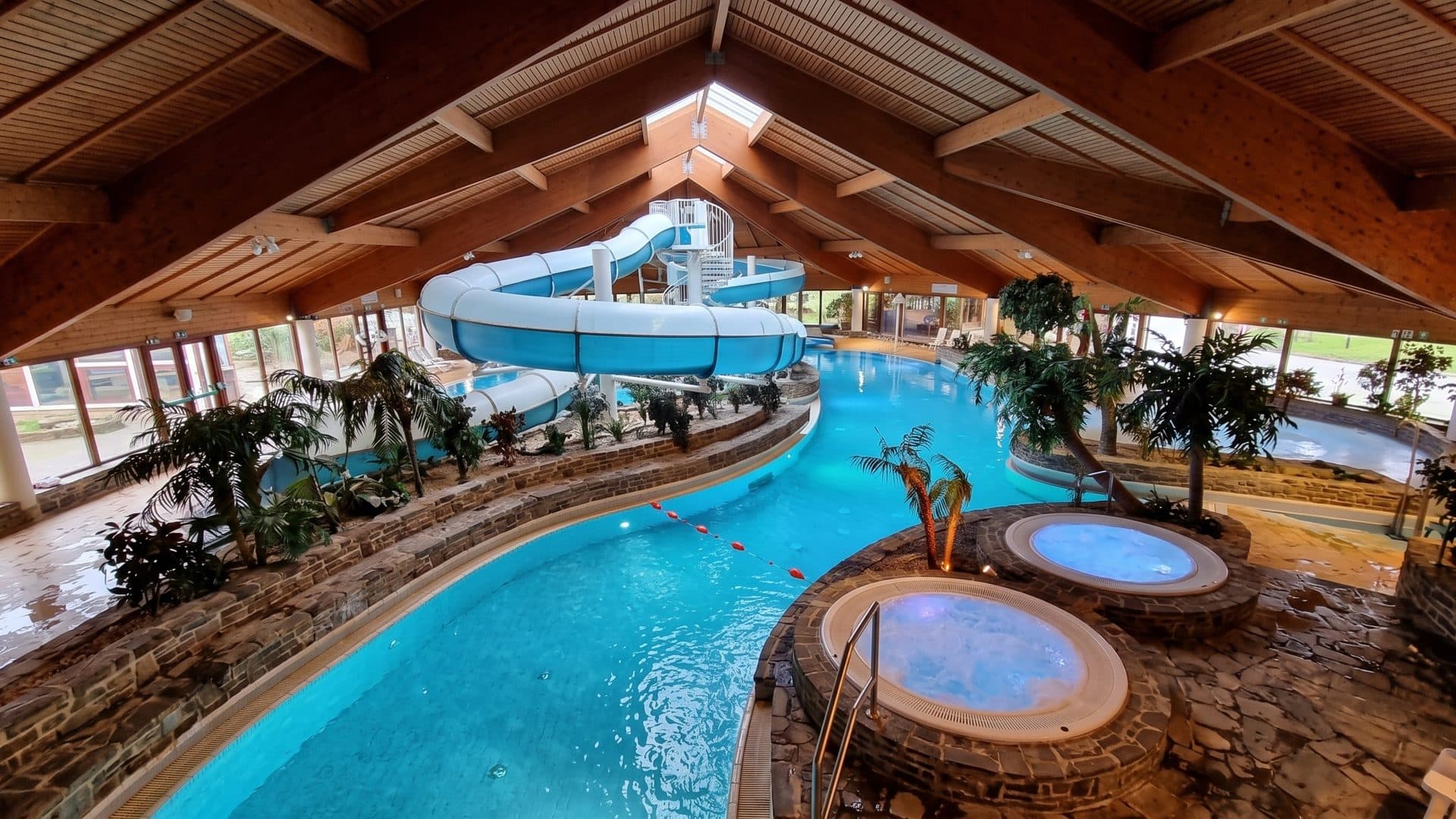 The winding indoor leisure pool of Hildorado with round whirlpools, palms and a white slide under a timber roof