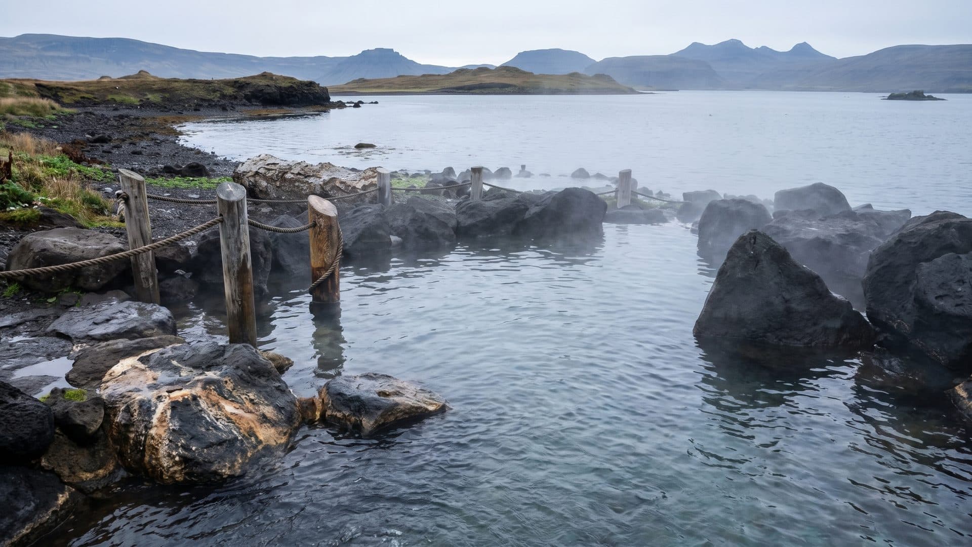 Hot pools among black rocks at the shoreline, steam drifting over the water towards the fjord.