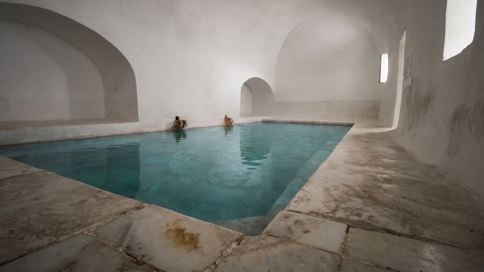 Vaulted stone bath chamber with green thermal water, daylight from a slit window high in the wall.
