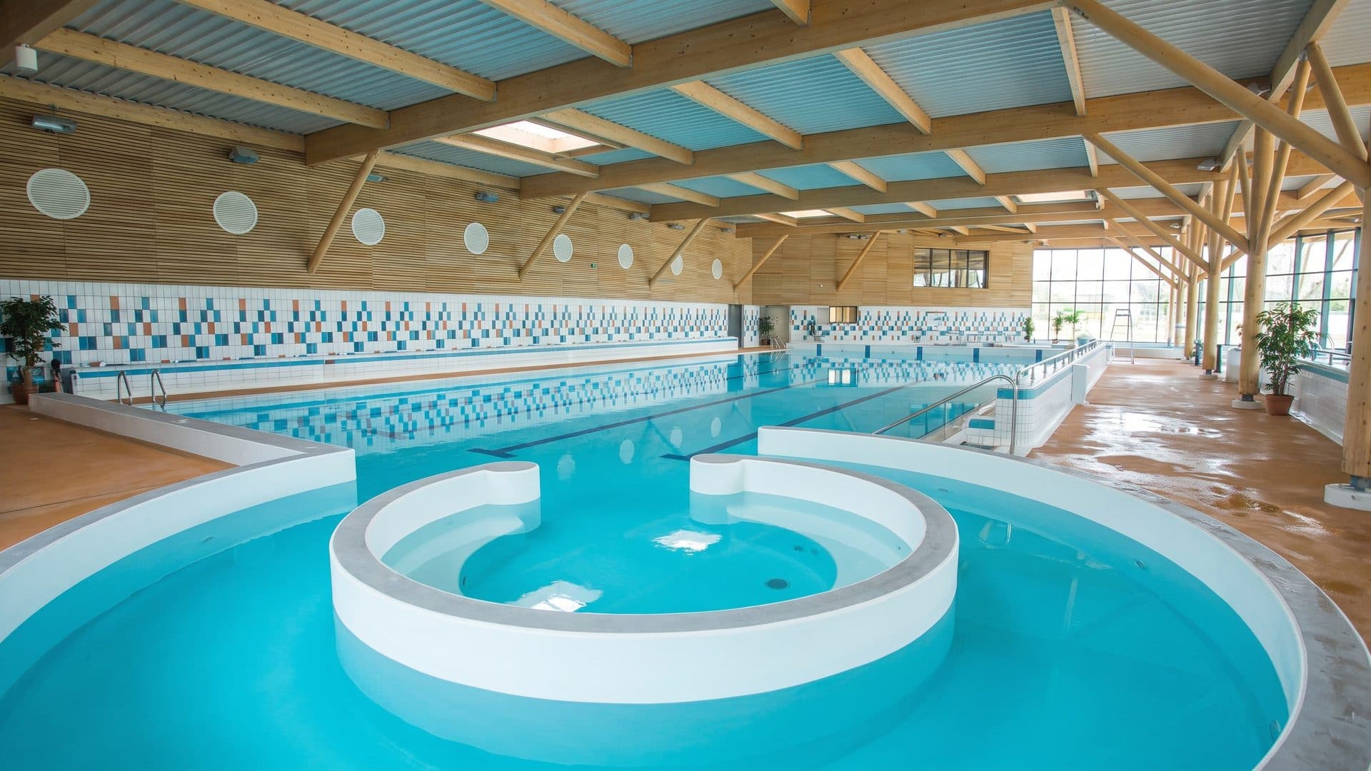 Indoor leisure pool with a round whirlpool island, in a hall with a timber roof and porthole windows