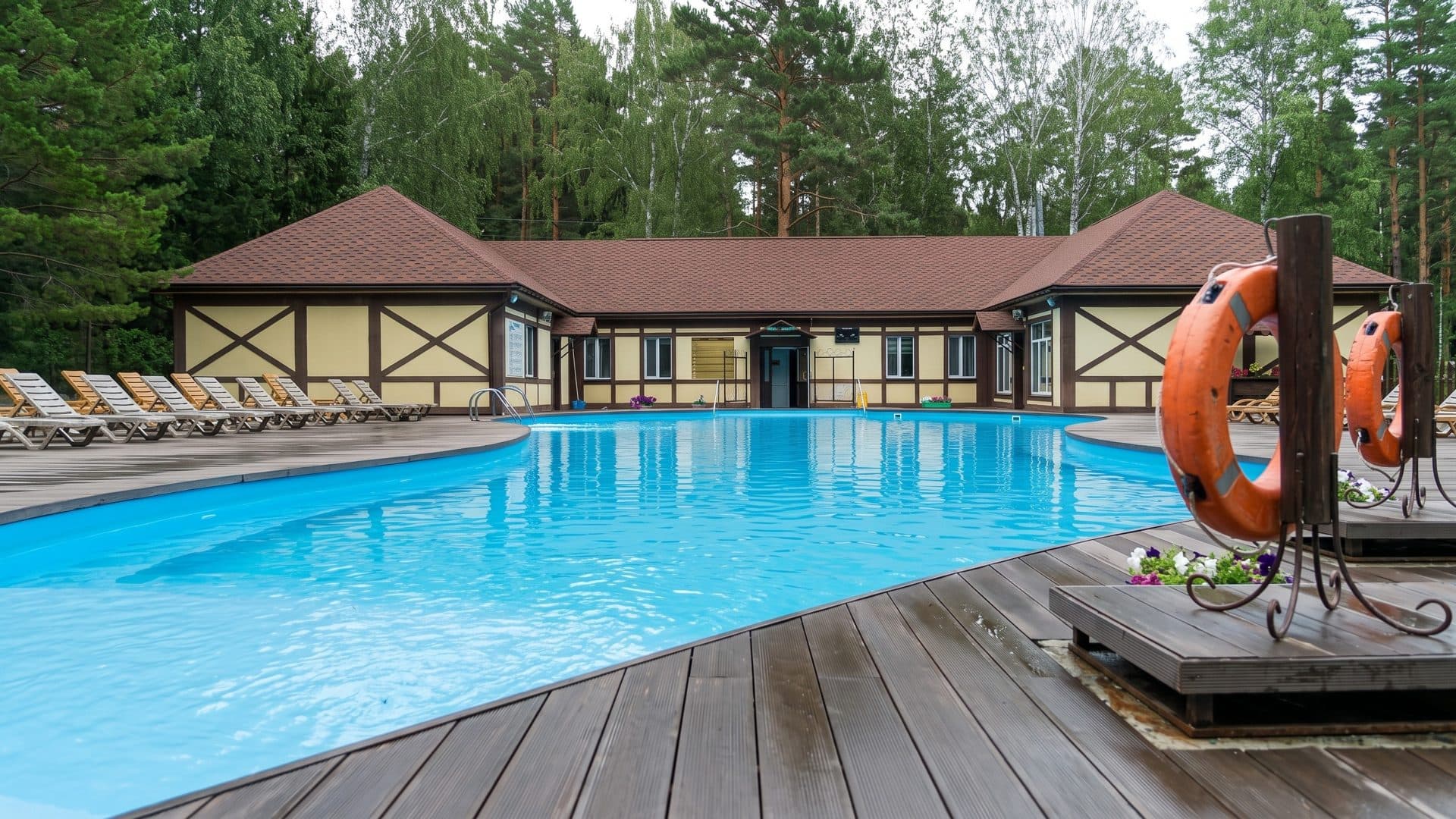 The curved outdoor pool of Ingala on a timber deck before the half-timbered spa building in a pine forest