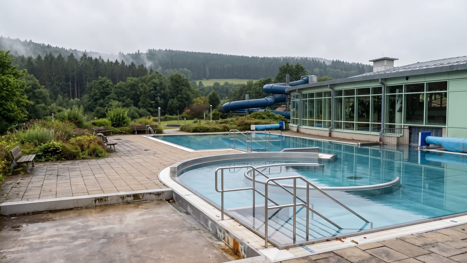 The outdoor pool of the Inselbergbad beside the glazed hall with its blue slide, wooded hills behind