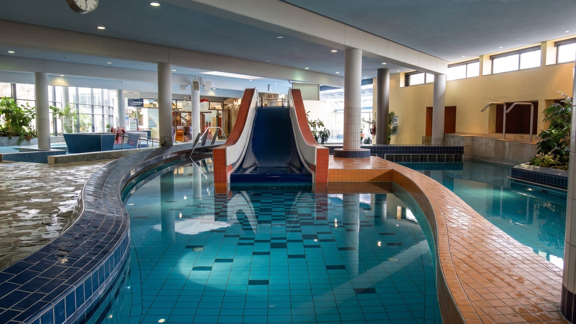 Indoor leisure pool with a winding channel, a red and blue slide and a curved tiled walkway
