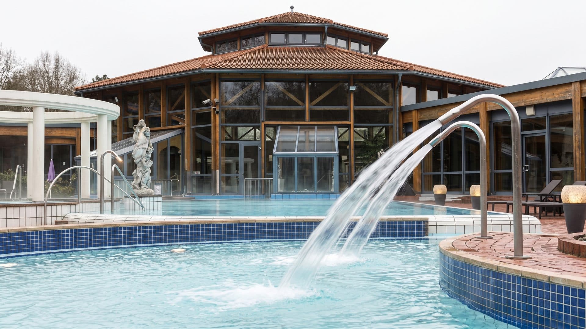 The outdoor brine pool of the Ith Sole Therme with arcing fountains playing across it and the timber-and-glass bath hall behind