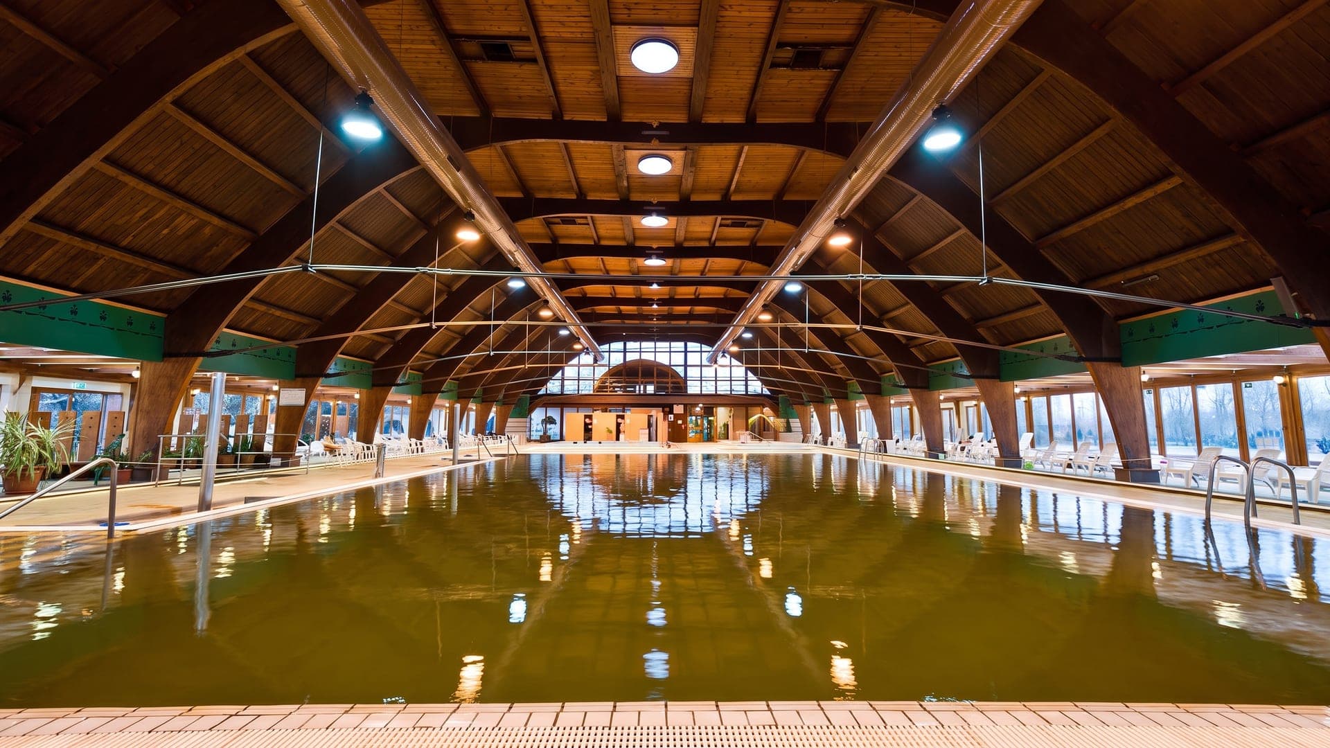 The indoor hall at Jonathermal in Kiskunmajsa, its brown thermal water under a wide timber-arched roof.