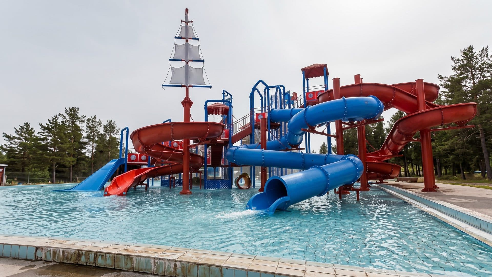 A pirate-ship slide tower with red and blue slides over the pool at Jukupark Kalajoki, pine trees around