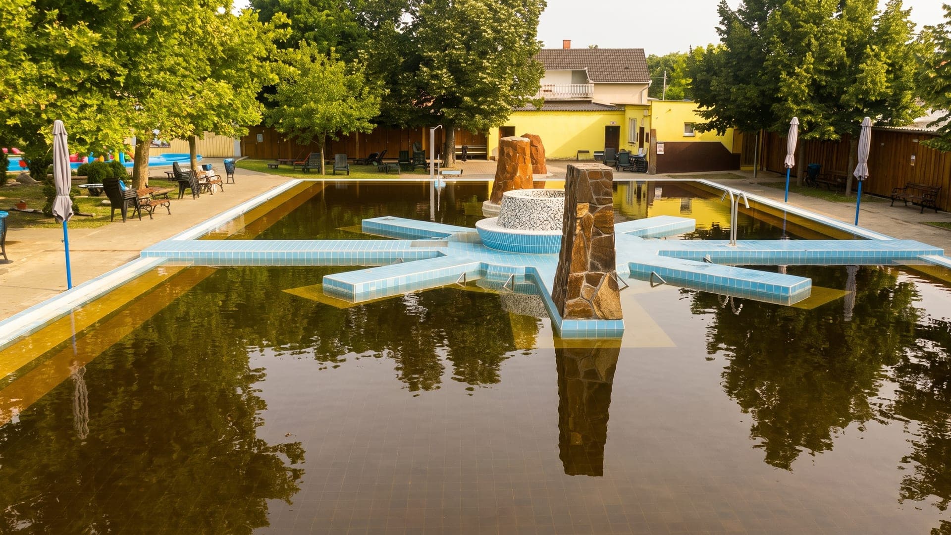 The outdoor thermal pool at the town bath in Kaba, its dark brown mineral water around tiled walkways and rock features.