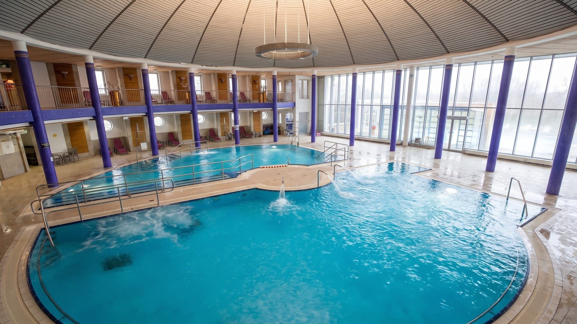Round indoor thermal pool under a domed ceiling, ringed by purple columns and a gallery of cabins