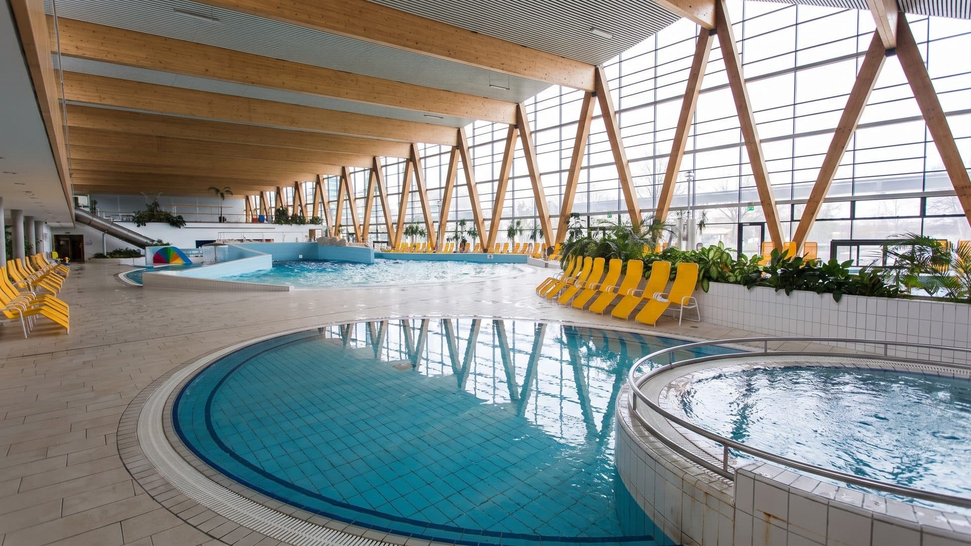 Indoor pools under a timber roof with triangular glazed trusses, and yellow loungers along the side