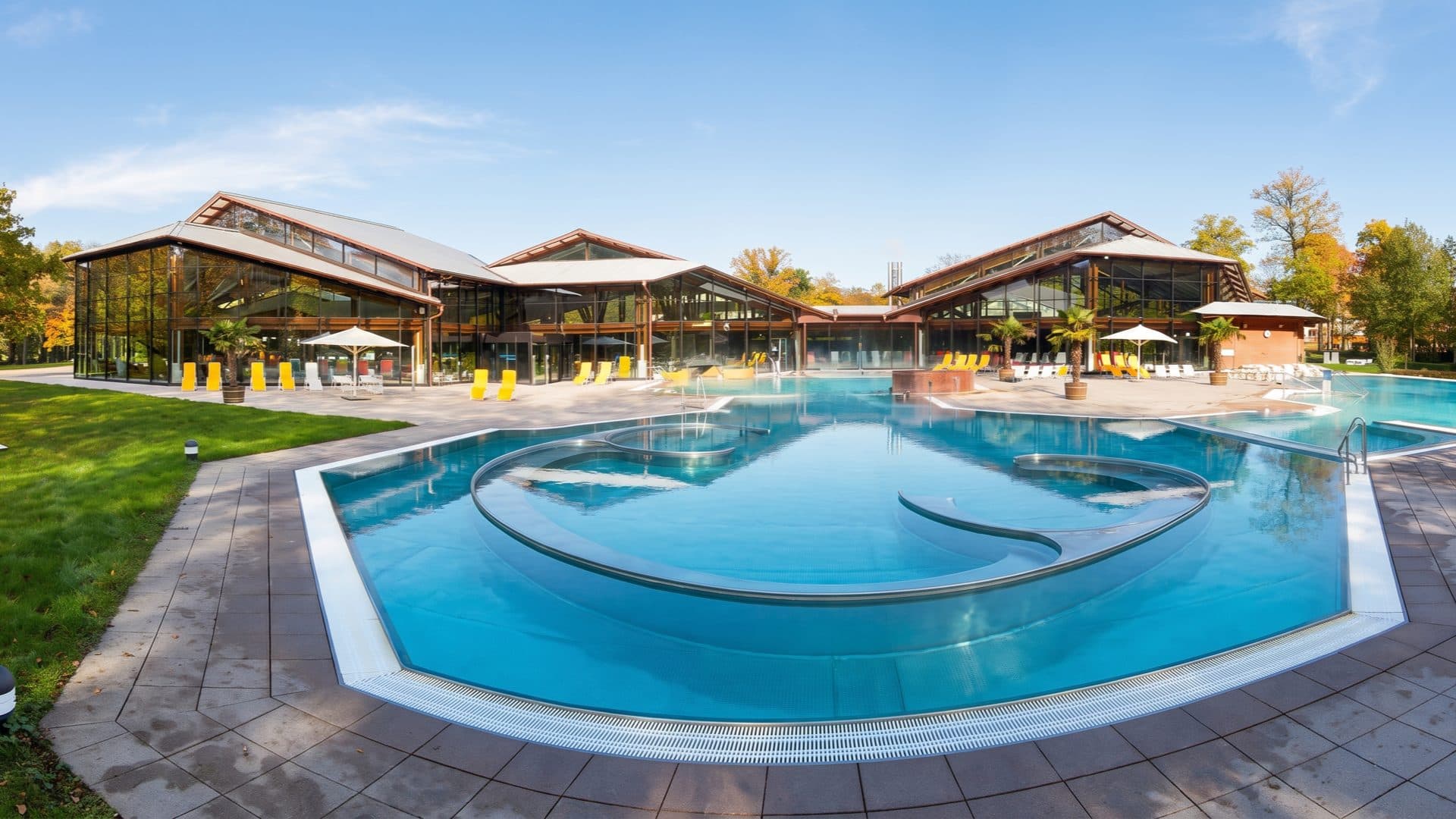 Curved outdoor thermal pool in front of three glass-and-timber pavilion halls, yellow loungers on the terrace and autumn trees around the lawn.