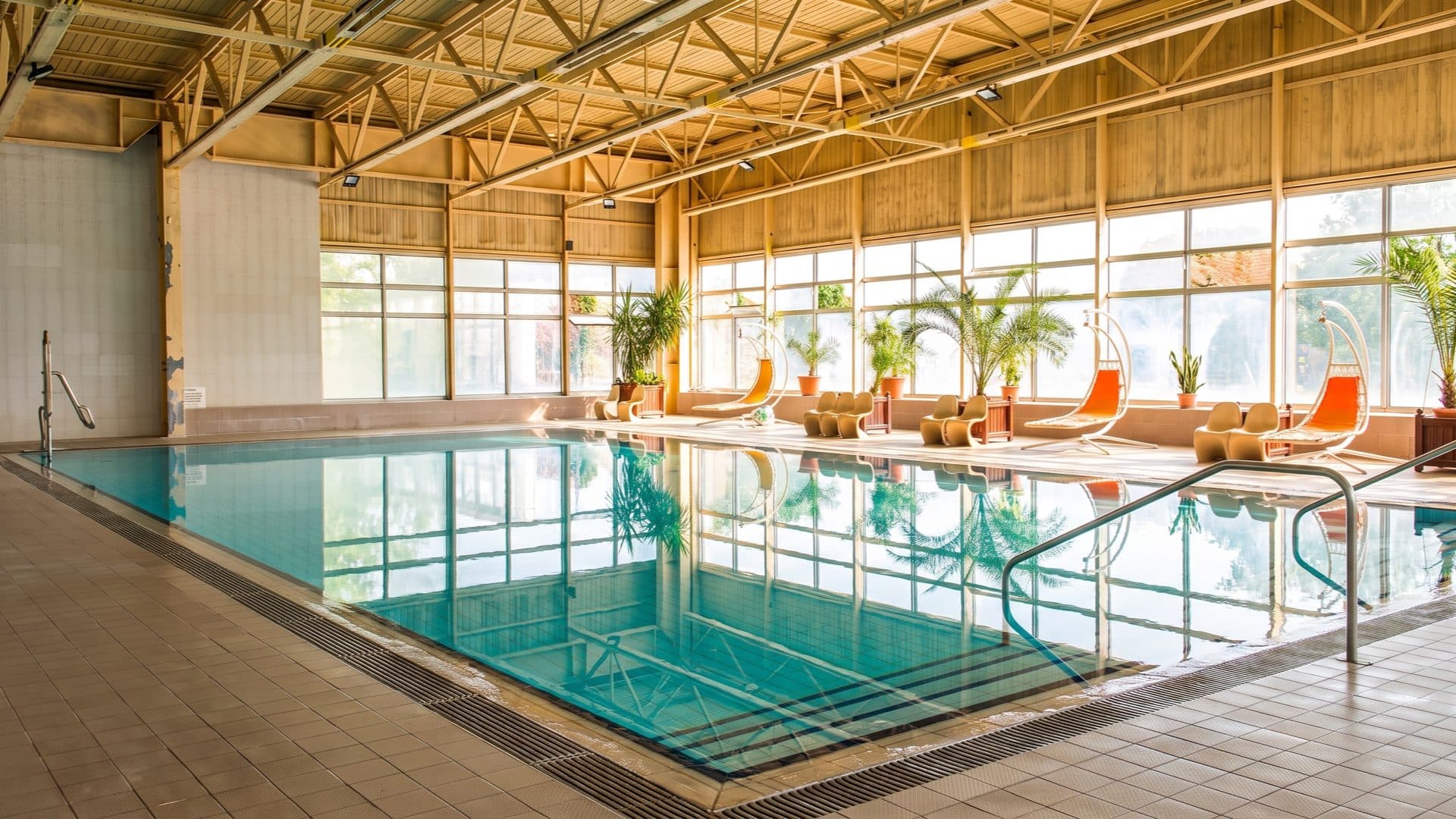 Indoor thermal pool in a bright hall with tall windows, potted palms and orange hanging pod chairs along the far side.