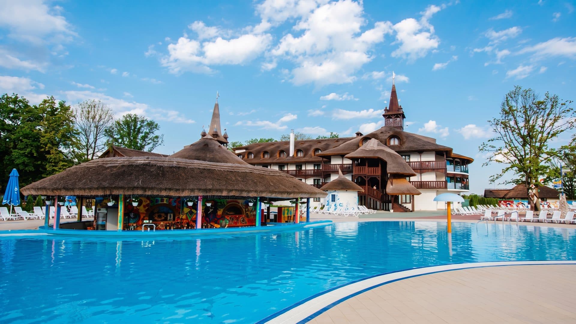 Large outdoor thermal pool with a thatched swim-up bar, and a folk-style resort building with steep thatched roofs and timber towers behind.