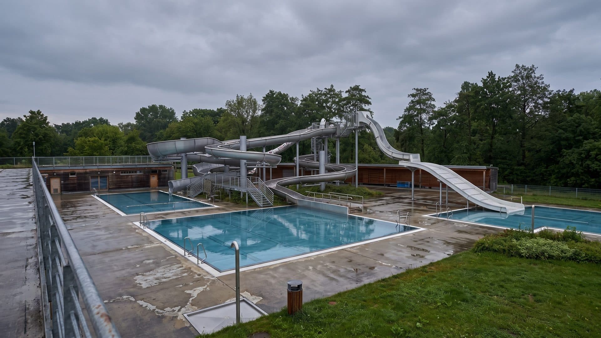 Outdoor pools and grey steel waterslides in a park, seen from a raised walkway on an overcast day