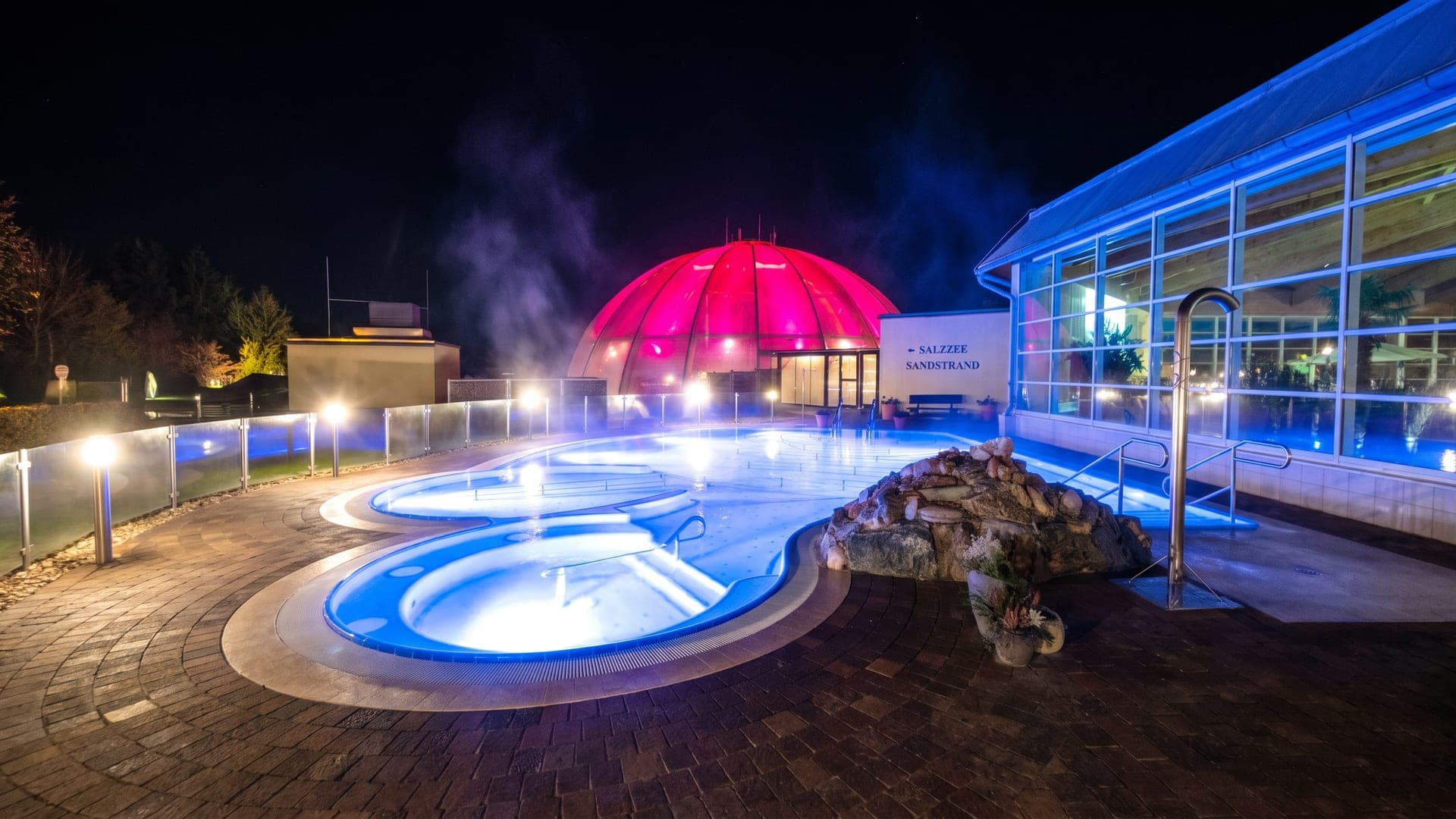 Outdoor pools lit blue at night beside the pink-lit dome