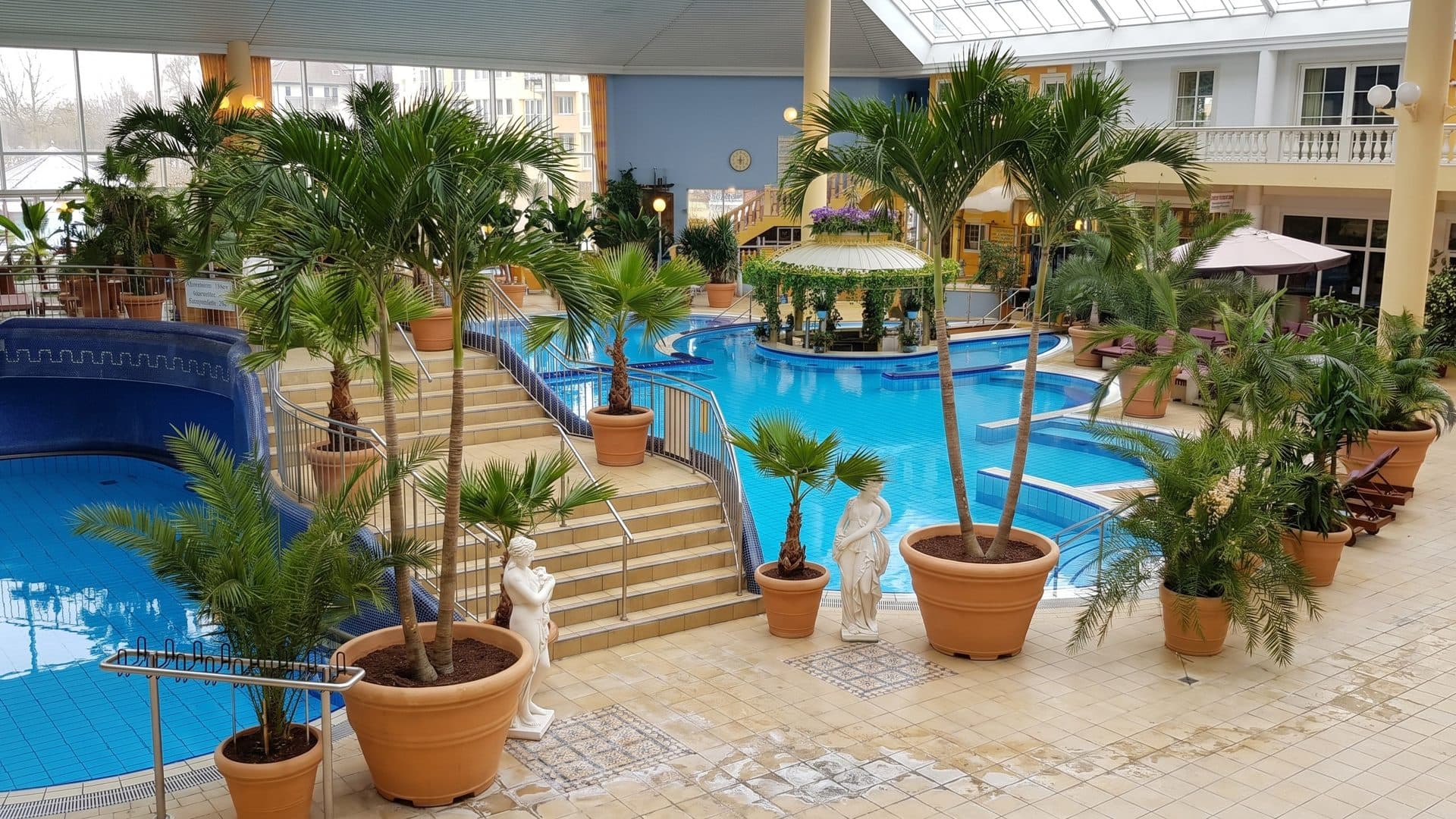 Indoor thermal pools with steps, potted palms and white statues in a light-filled hall