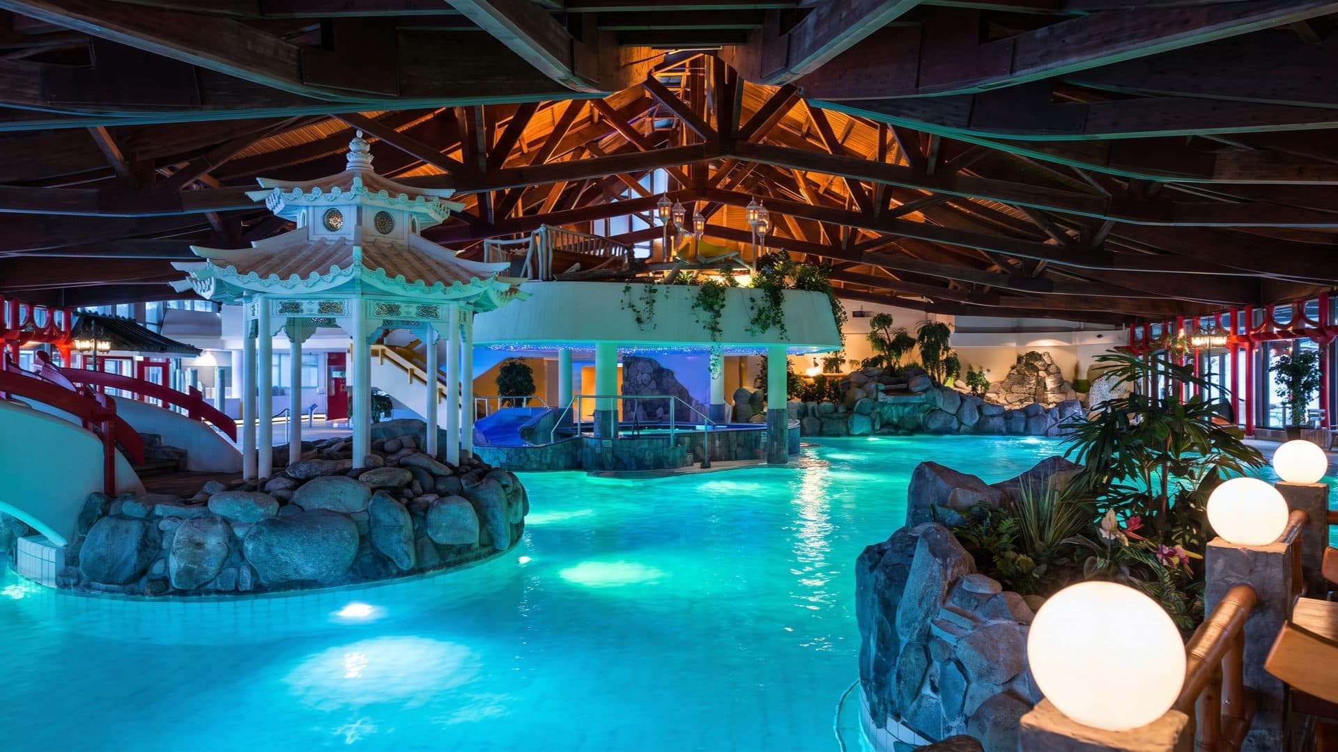 Indoor thermal pool at night with an Asian-themed white pagoda and a red bridge, rockwork and glowing globe lamps reflected in turquoise water.