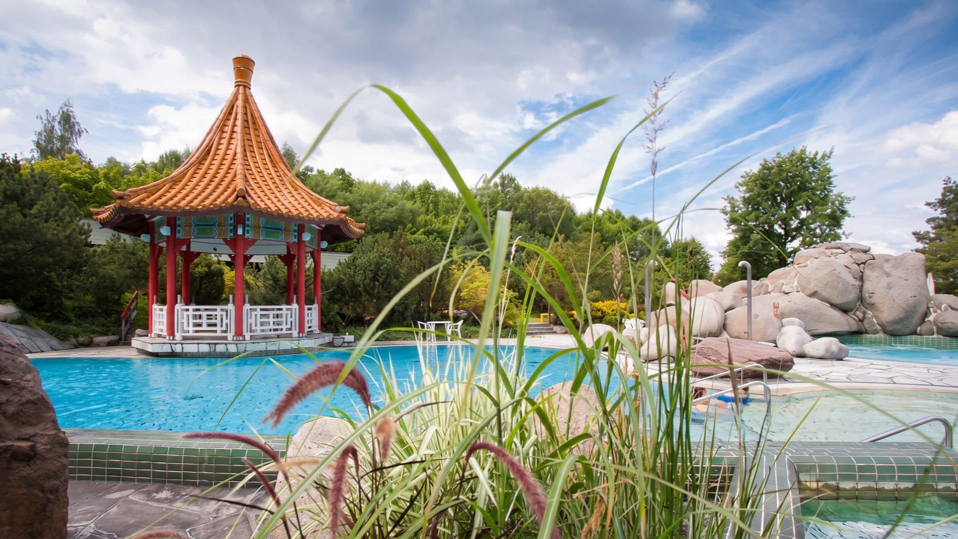 Outdoor thermal pool beside an ornate Chinese-style pagoda pavilion, framed by boulders and tall ornamental grasses.