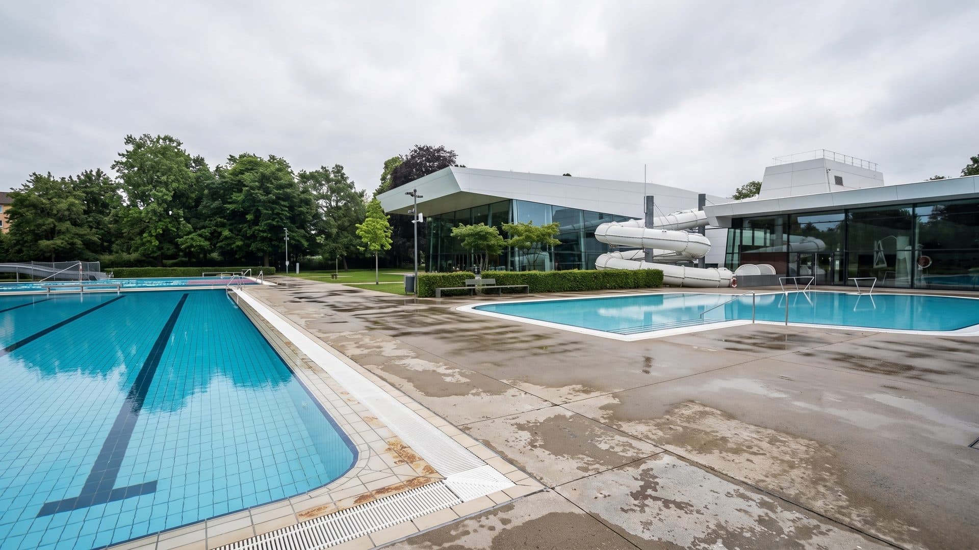 Outdoor lap pool and leisure pool on a wide deck, with the white pool building and its slide behind