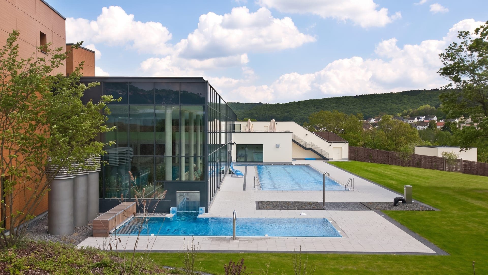 The outdoor pools at the Lahn-Dill-Bergland-Therme in Bad Endbach, beside the glass building with the wooded hills of the Bergland behind.