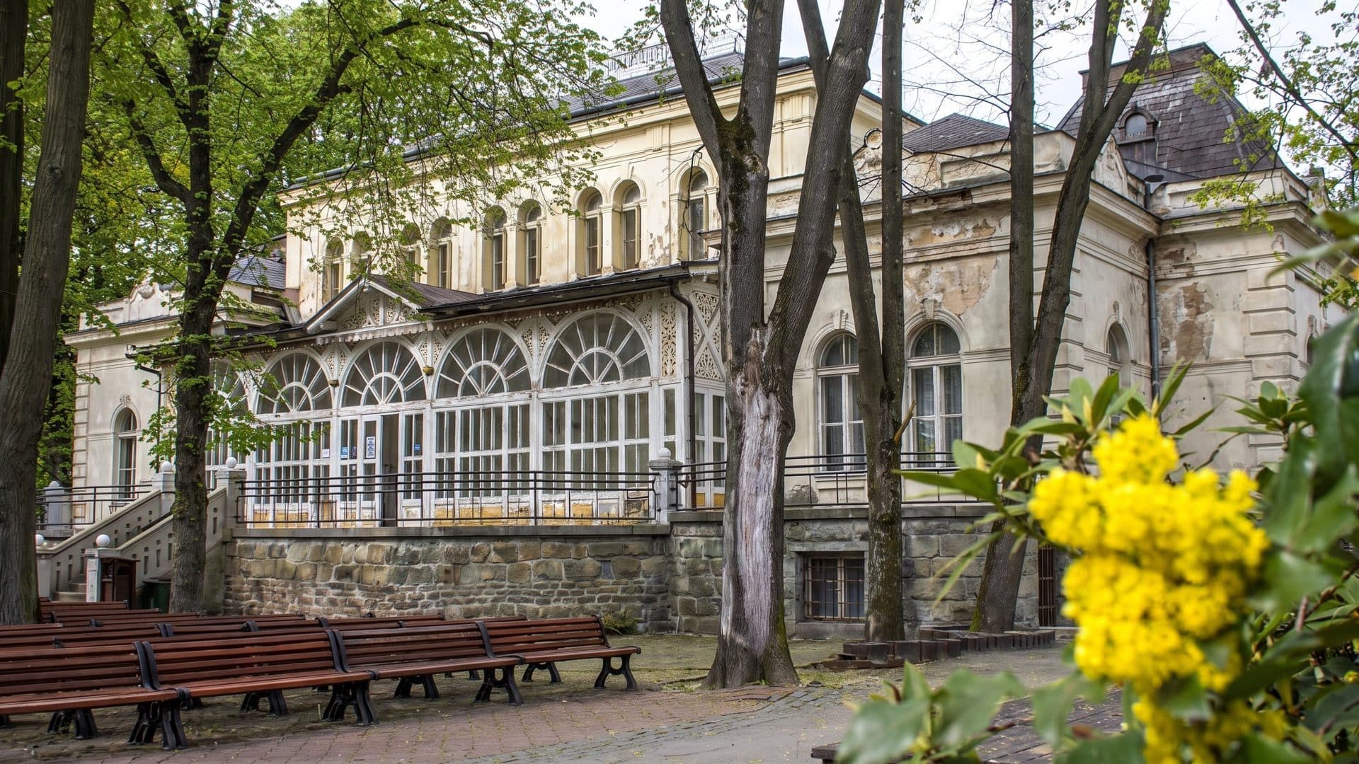 The historic spa building with its arched glass veranda, seen through trees past park benches and a flowering shrub