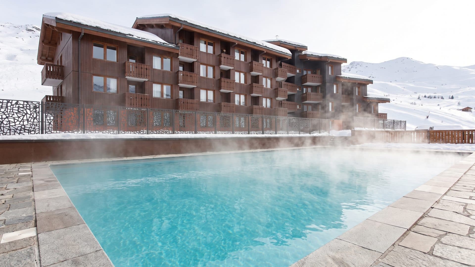 The steaming outdoor pool of Les Bains de Belle Plagne below the timber resort building in the snow