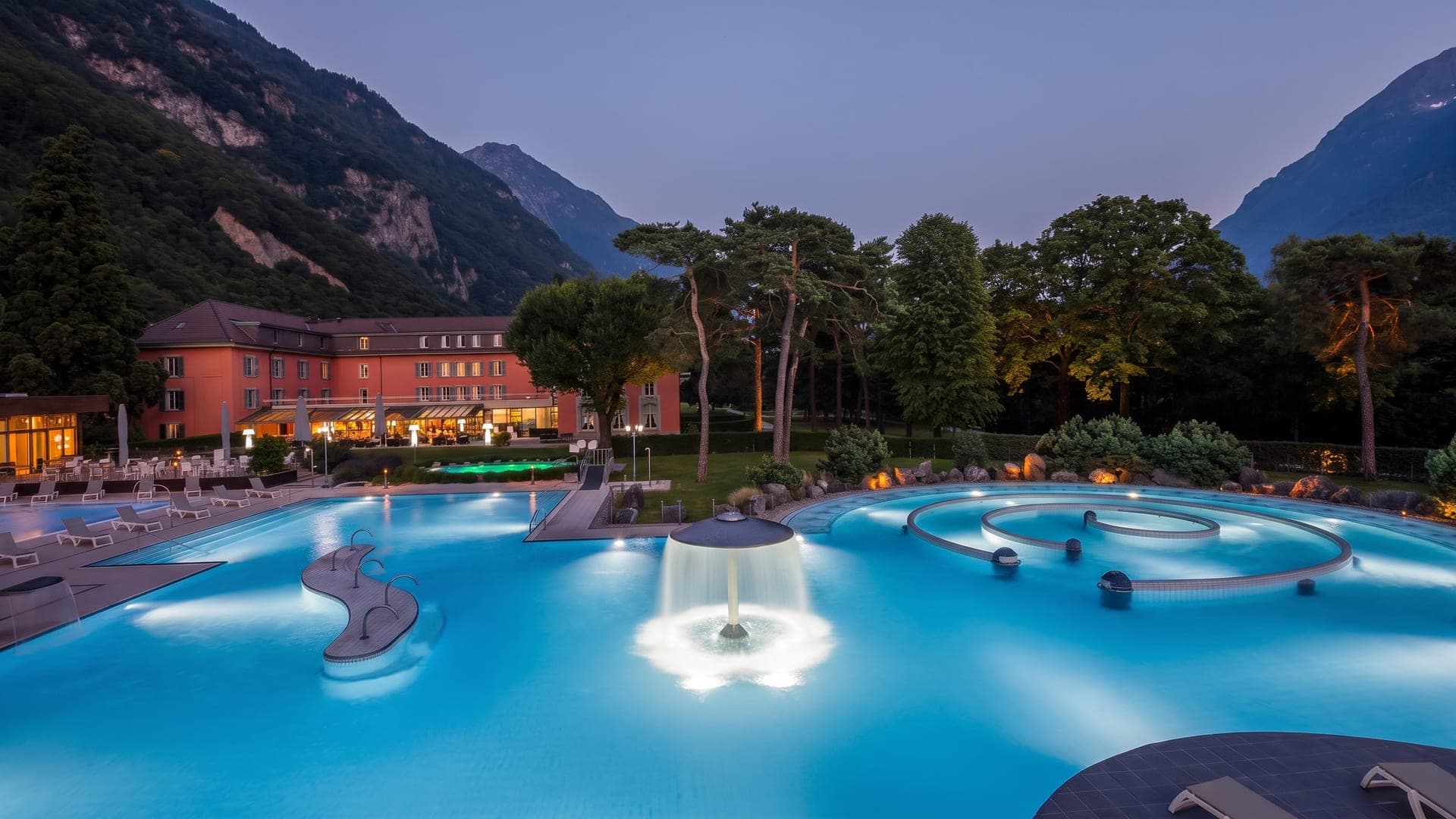 Illuminated outdoor pools at dusk, with mountains and the pink hotel behind