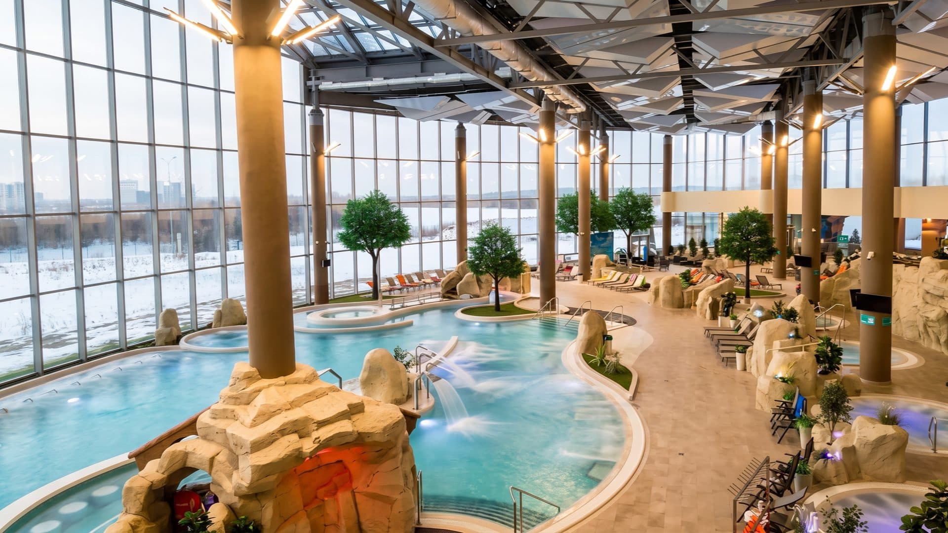 Large modern indoor thermal water-park with curved pools and artificial rockwork, tall glass walls looking out onto a snowy landscape.