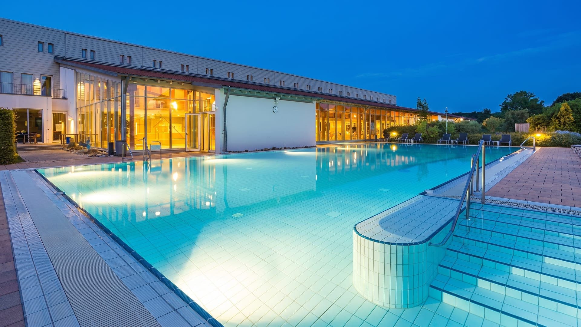The outdoor pool at the Lohengrin Therme in Bayreuth at dusk, the lit building reflected in the water.