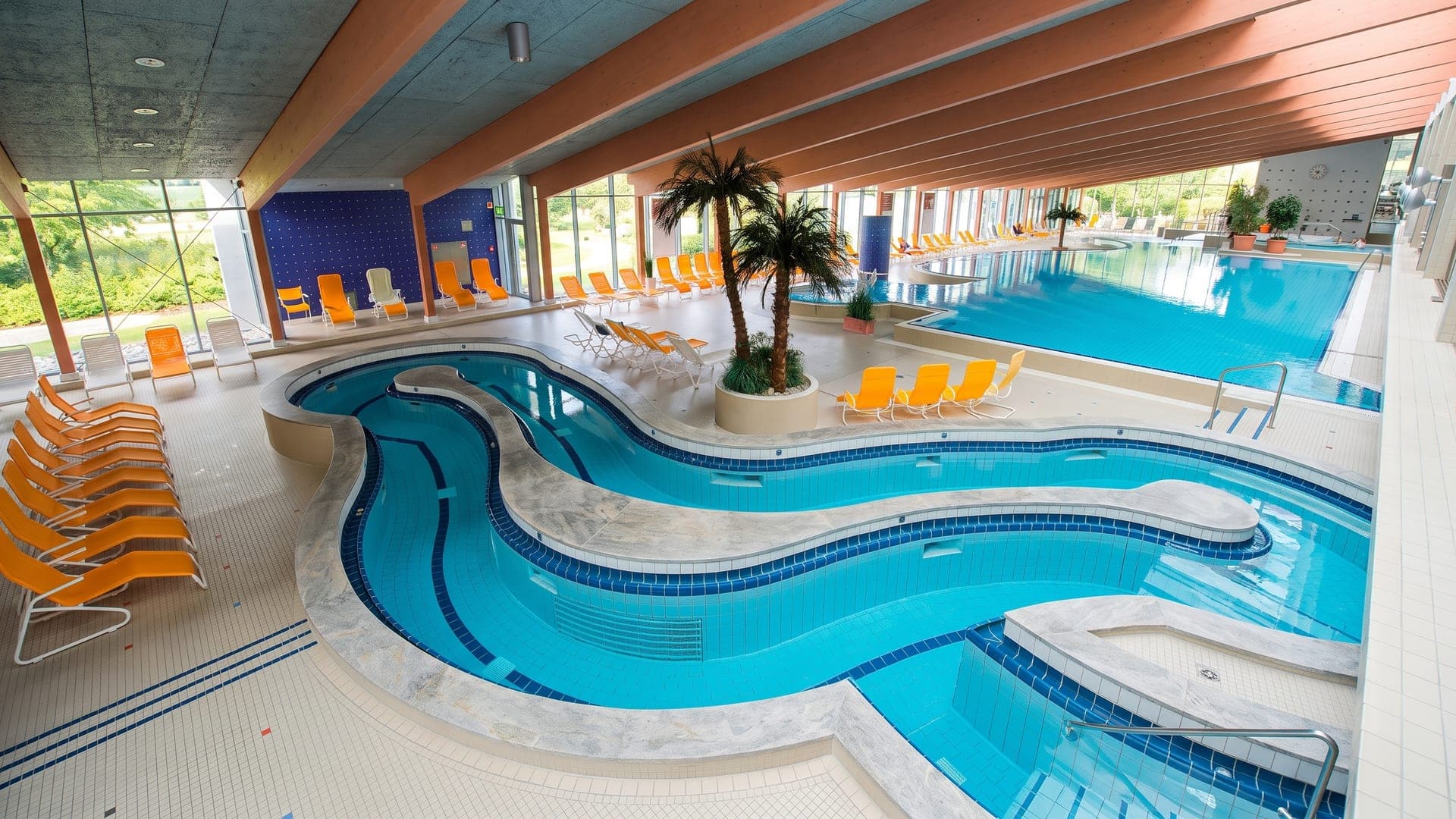 The indoor pool at the Lohengrin Therme in Bayreuth, a winding channel under a timber ceiling with palms and orange loungers.