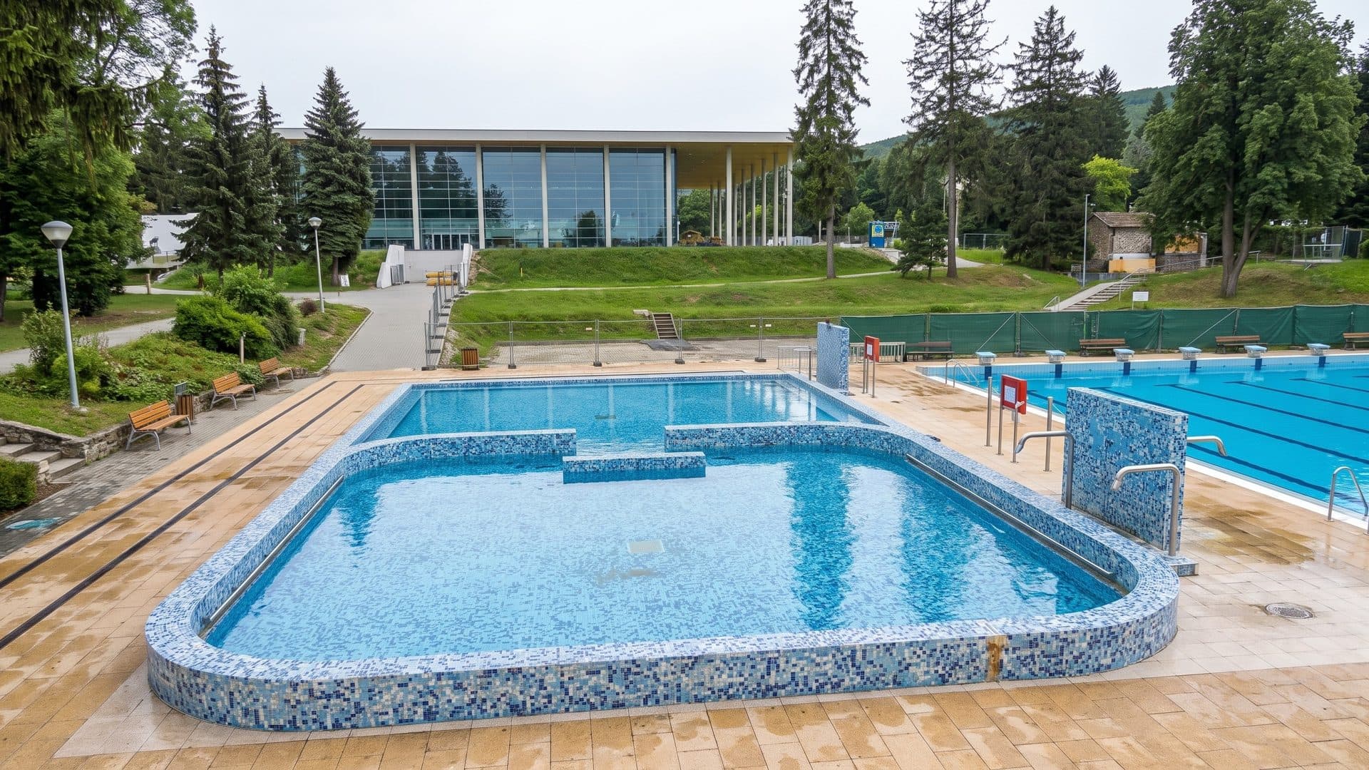 Mosaic-tiled outdoor thermal pool beside the sports pool, with the glazed bath house among spruce trees