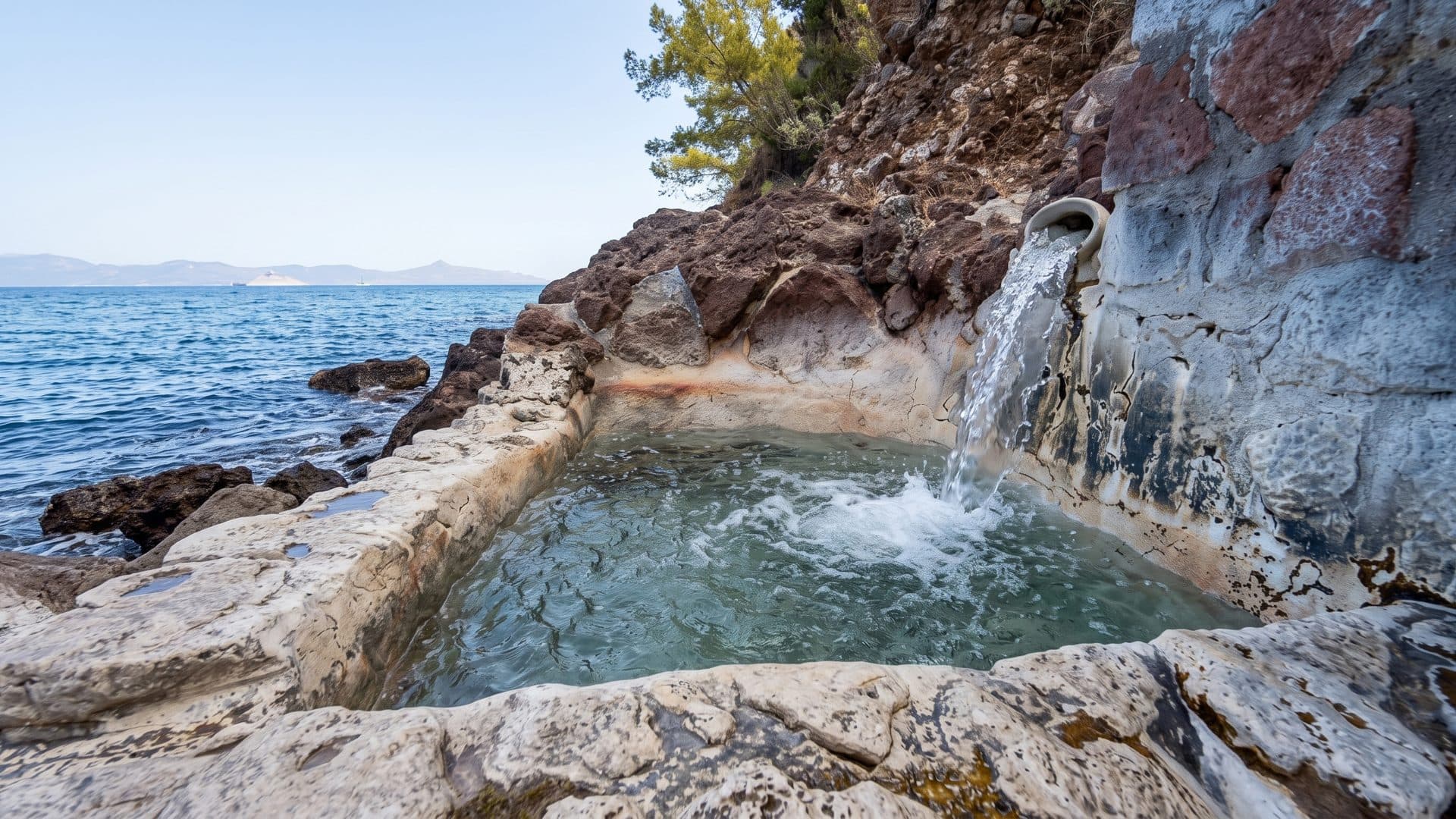 Rock-cut thermal basin on the shore, water running into it from the cliff, the sea and the headland behind.