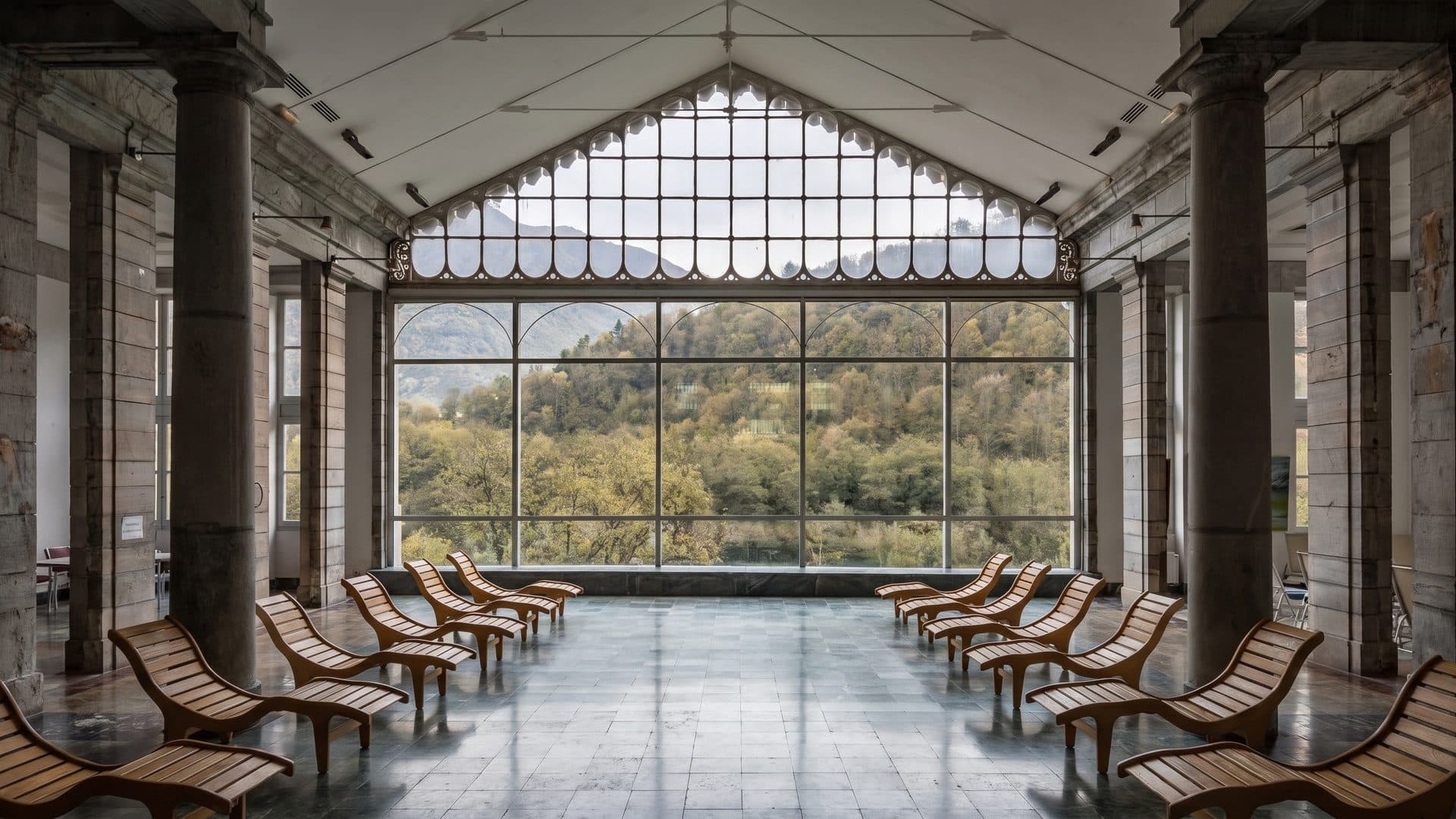 The colonnaded relaxation hall at Luzea, wooden loungers in rows on a marble floor before a tall window wall onto the woods