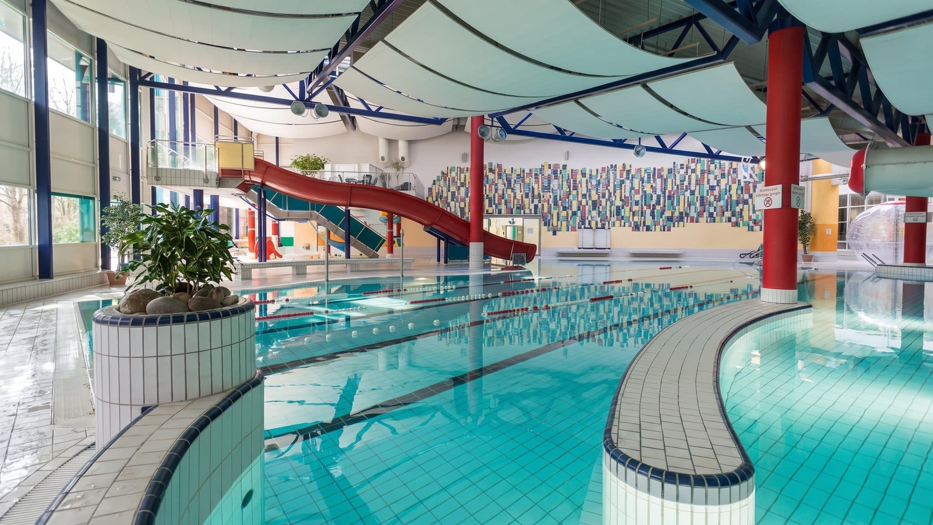 Indoor pool hall with a curved tiled bench, a red slide and white sail-like ceiling panels