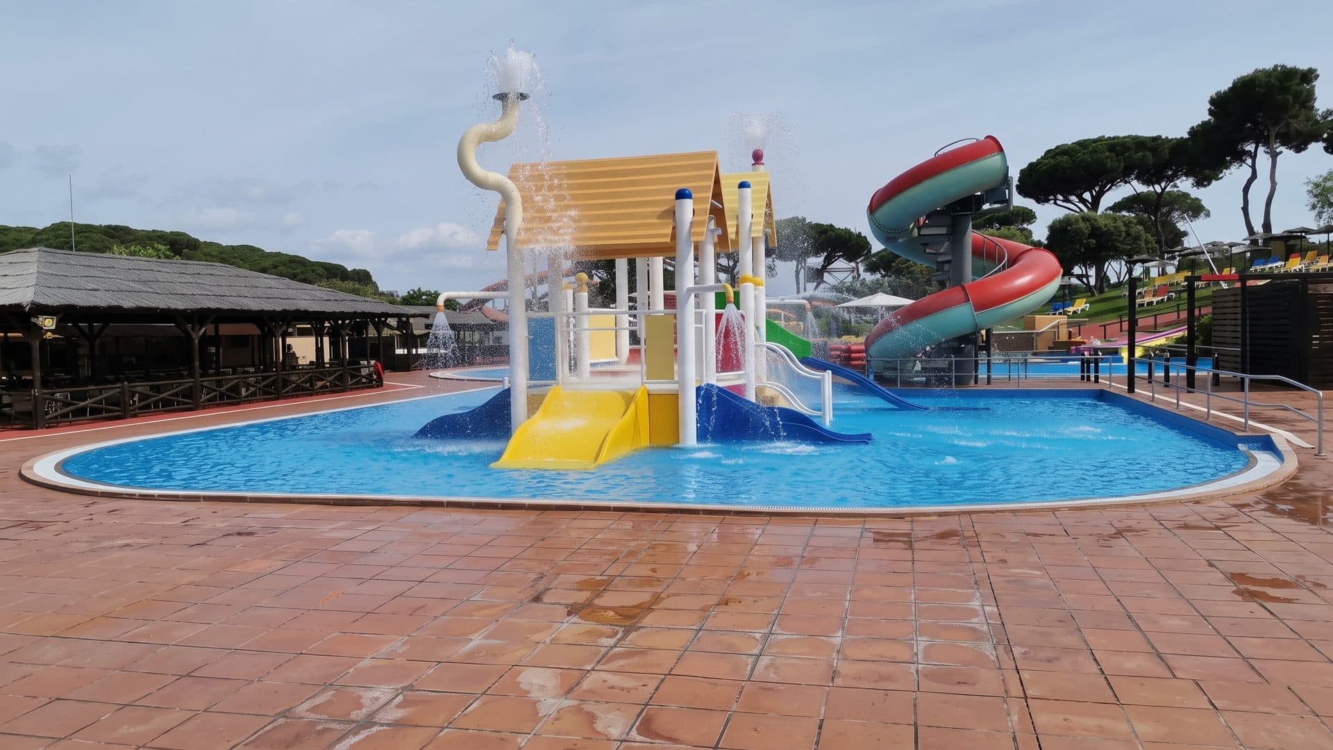 A children's pool at Marineland Catalunya with a yellow play house, red spiral slides and pine trees behind