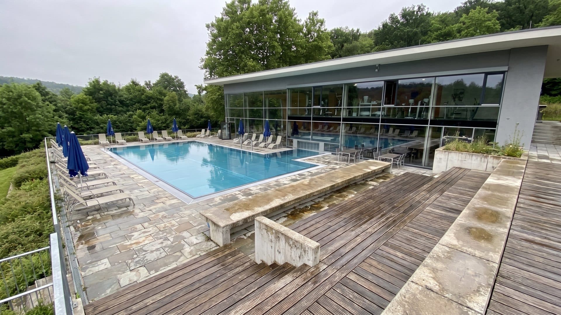 Outdoor thermal pool below a timber sun deck, with the glazed bath house on a green hillside