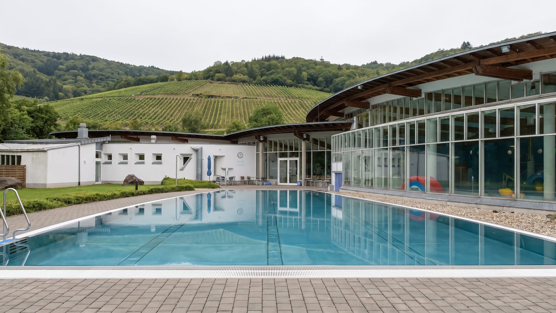 The outdoor pool of Mosel-Therme with the curved glazed hall behind it and vineyard slopes above