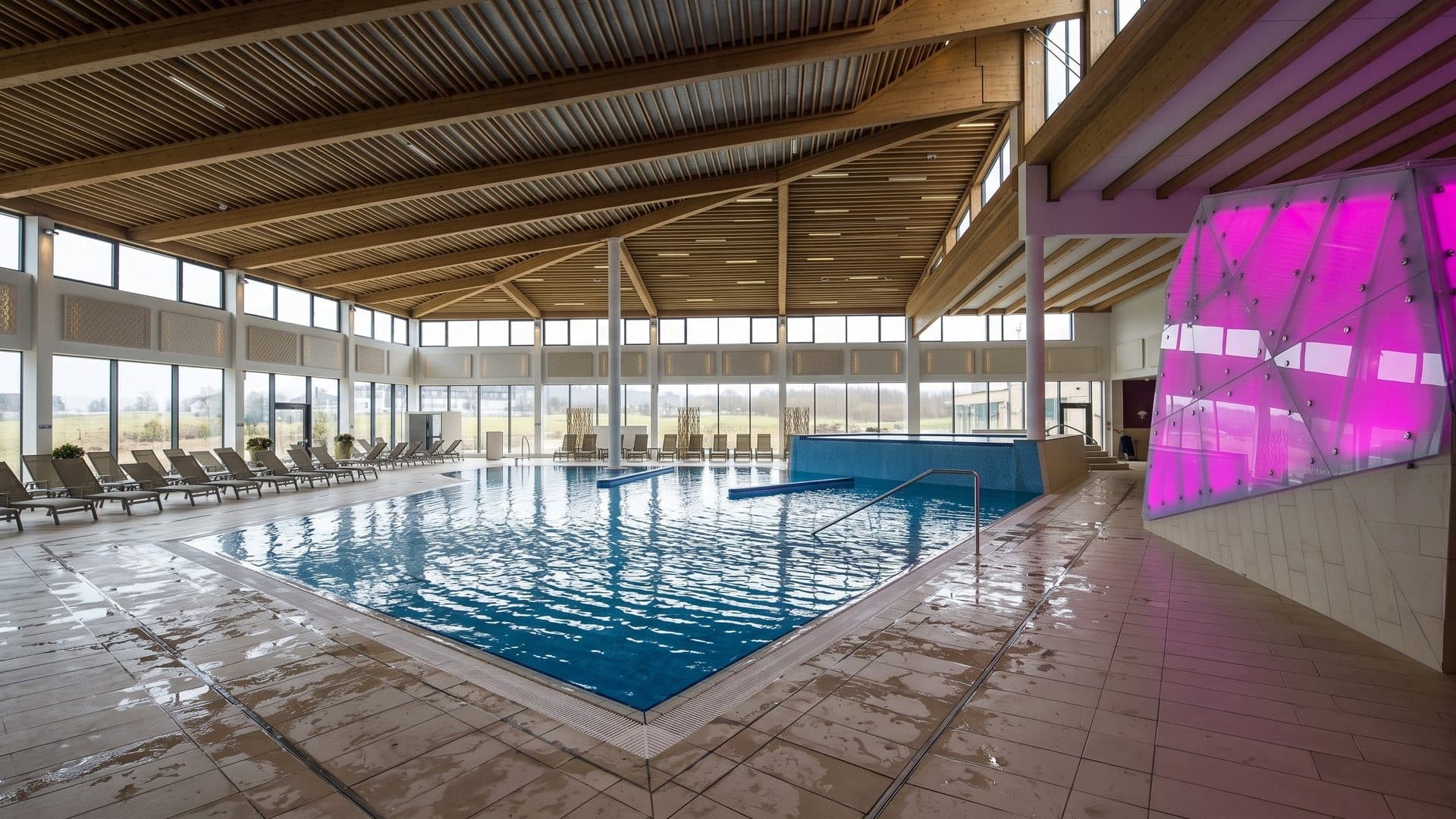 The indoor pool of the Narzissentherme in Bad Aussee under a timber ceiling, with a glowing pink glass wall