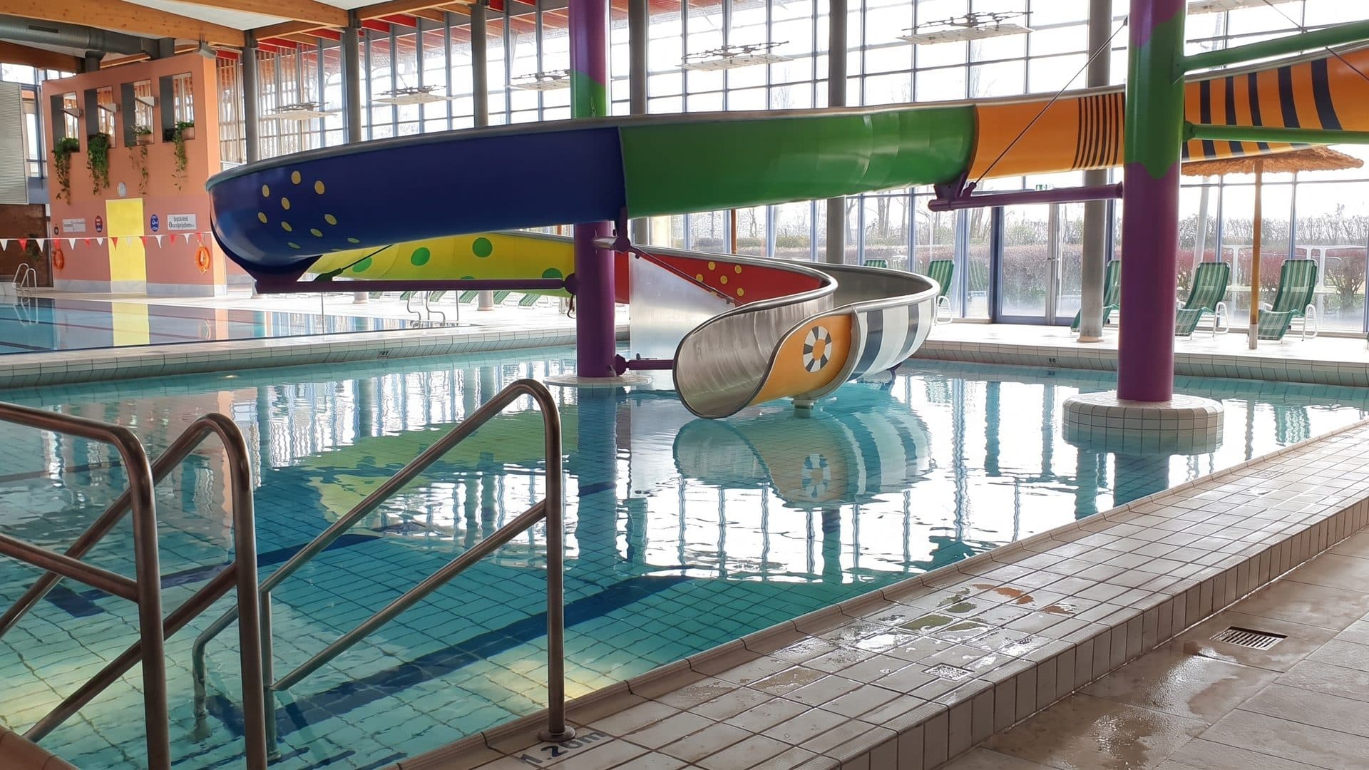 Indoor pool with steps and handrails beneath a colourful tube slide on purple and green columns
