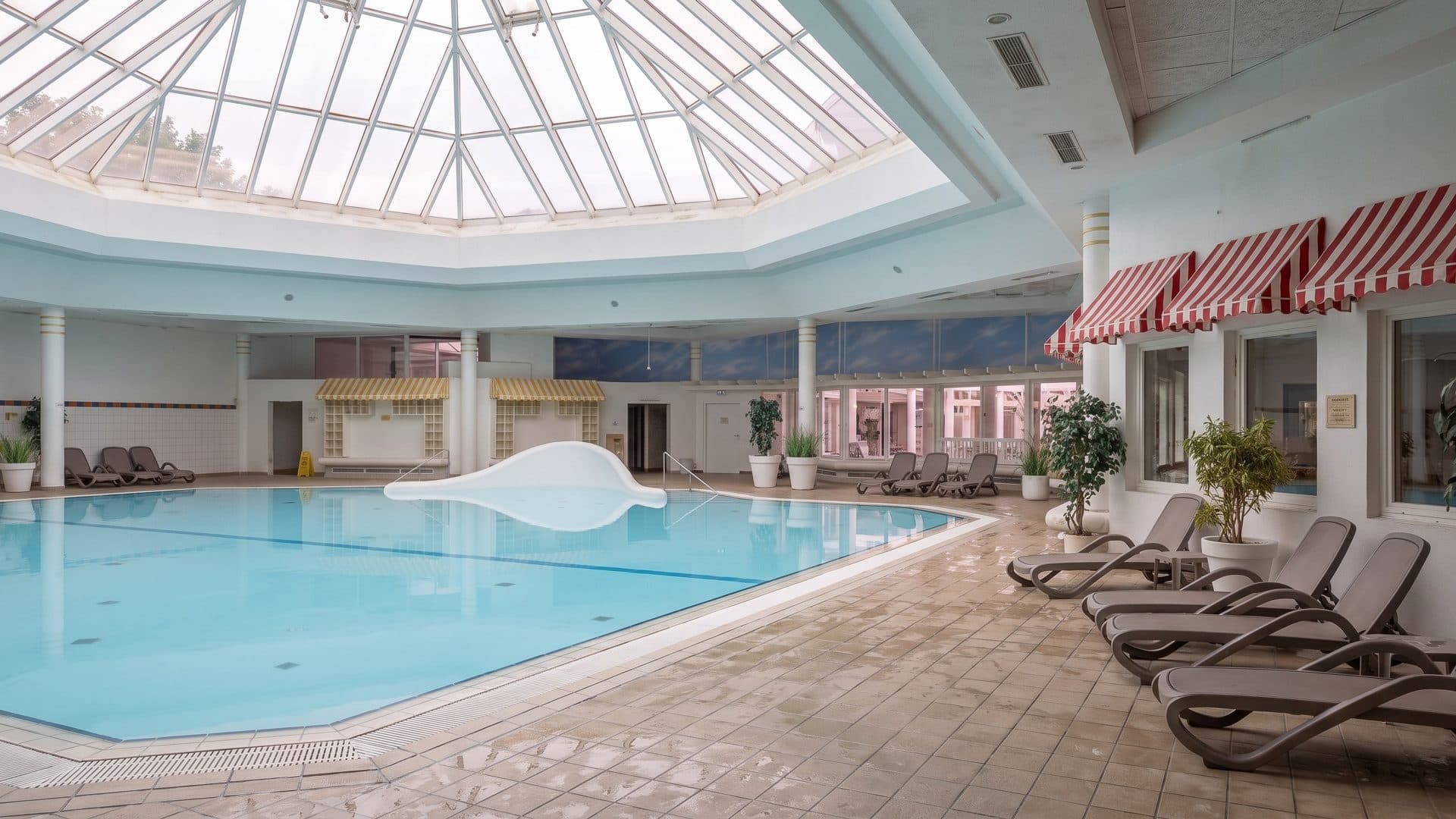 Indoor pool under an octagonal glass skylight, with striped awnings and loungers along the side