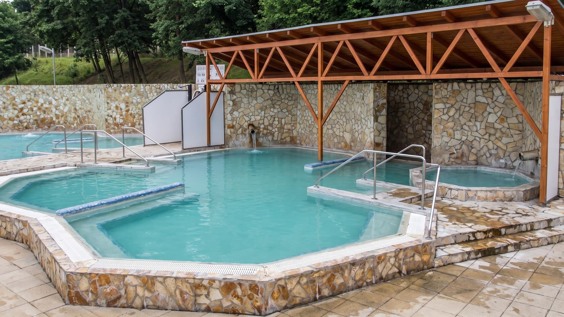 Outdoor thermal pool under a timber-framed roof, with a stone wall and a smaller whirlpool beside it