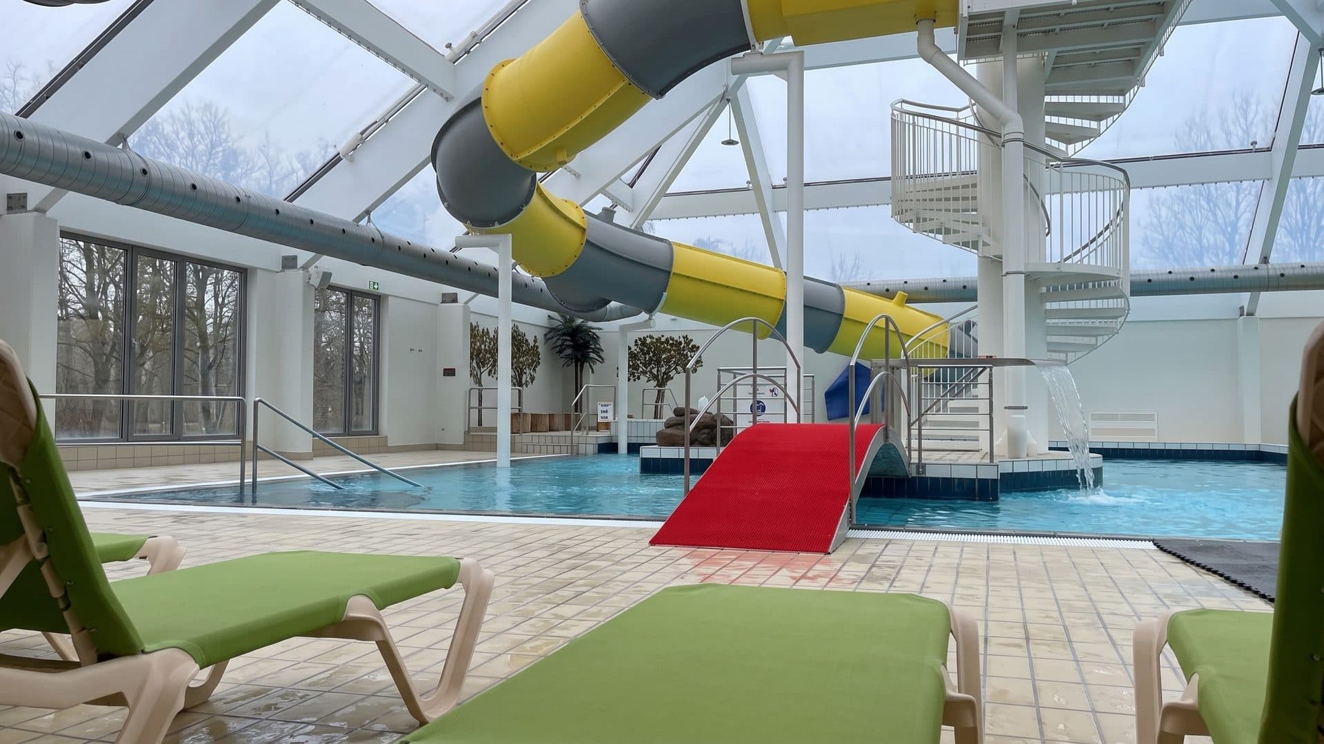 A yellow and grey tube slide under the glass roof of the Nyborg swimming hall, green loungers beside the pool