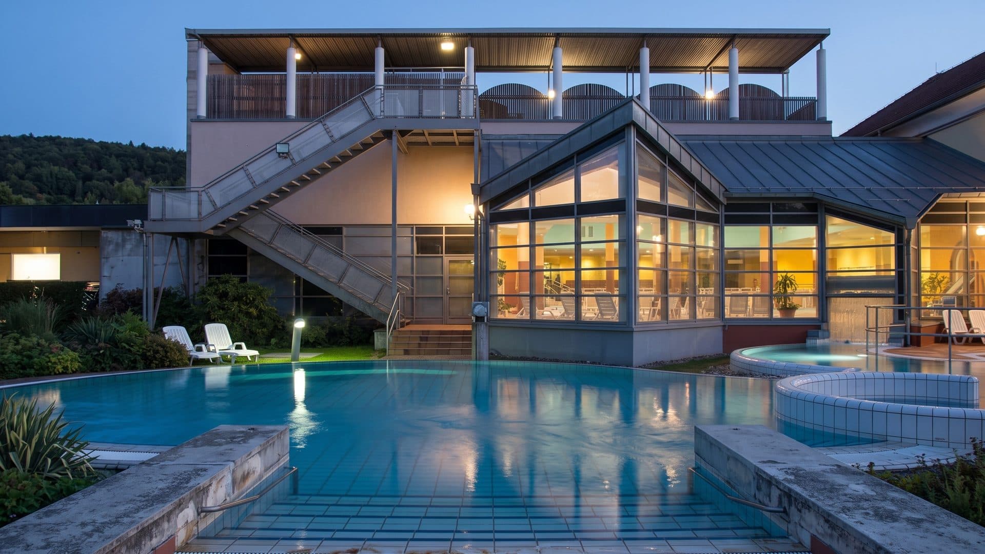 The outdoor thermal pool of the Odenwald-Therme at dusk in front of the lit glazed bath hall and its outside staircase