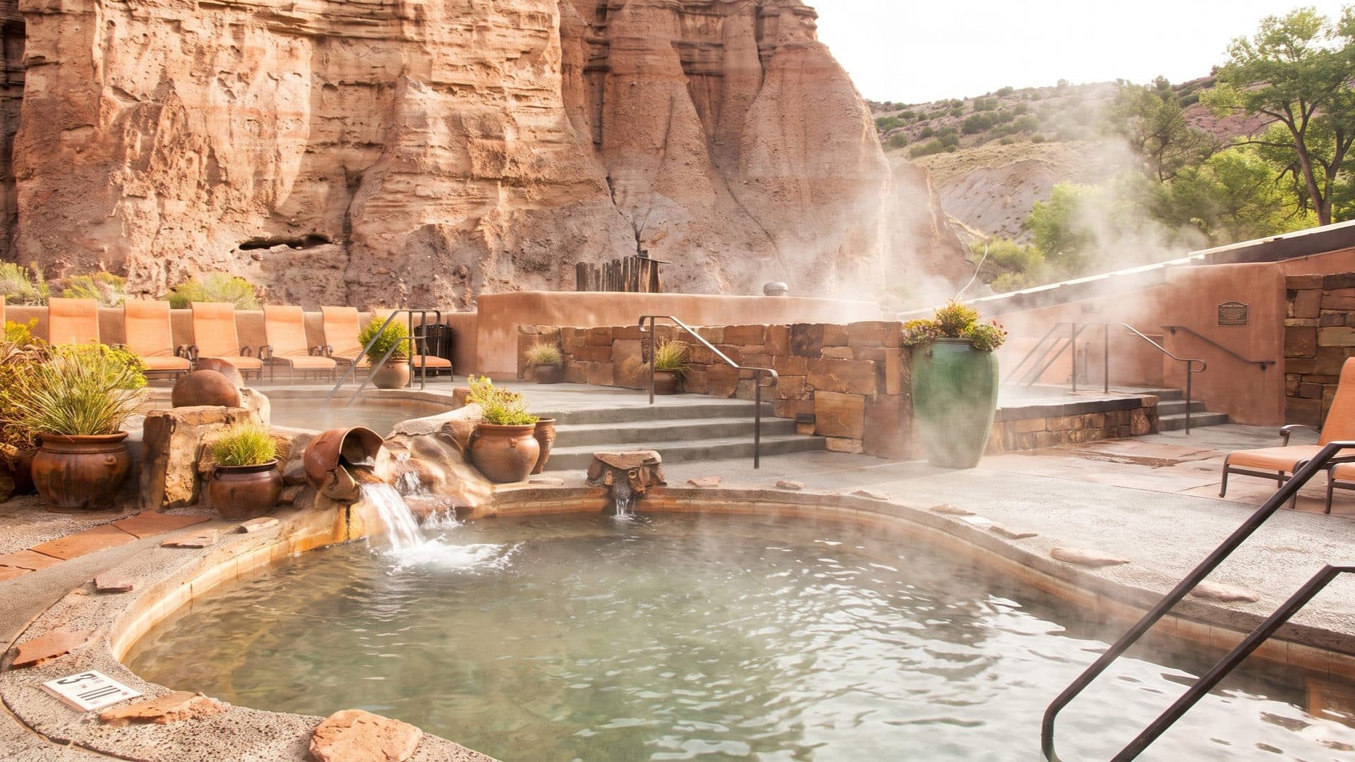 Steaming round mineral pool at the foot of tan sandstone cliffs, with adobe walls and terracotta pots around a desert spa terrace.