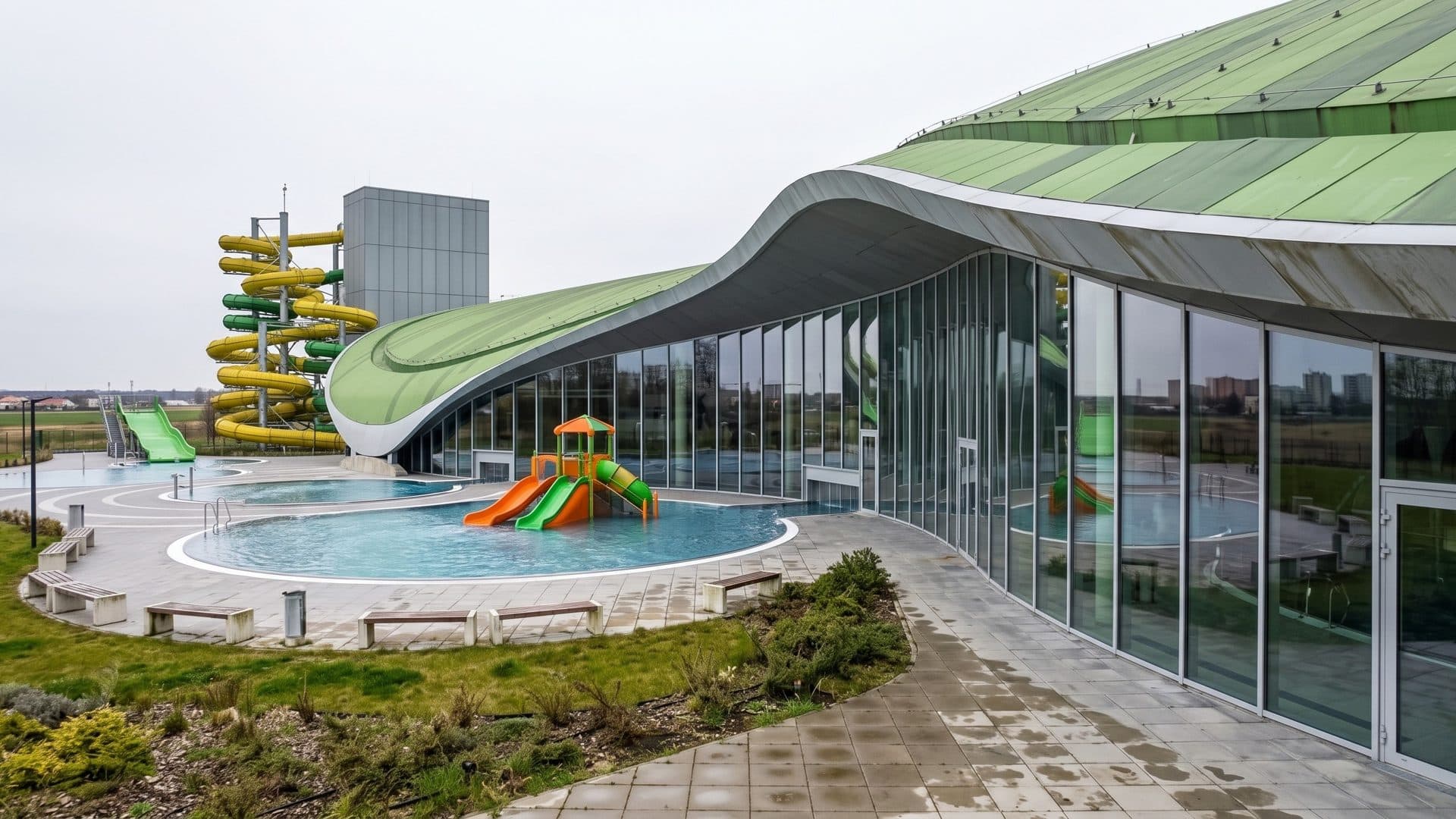 The outdoor pools of Park Wodny Koszalin beside the glazed hall with its green wave-shaped roof and slide tower