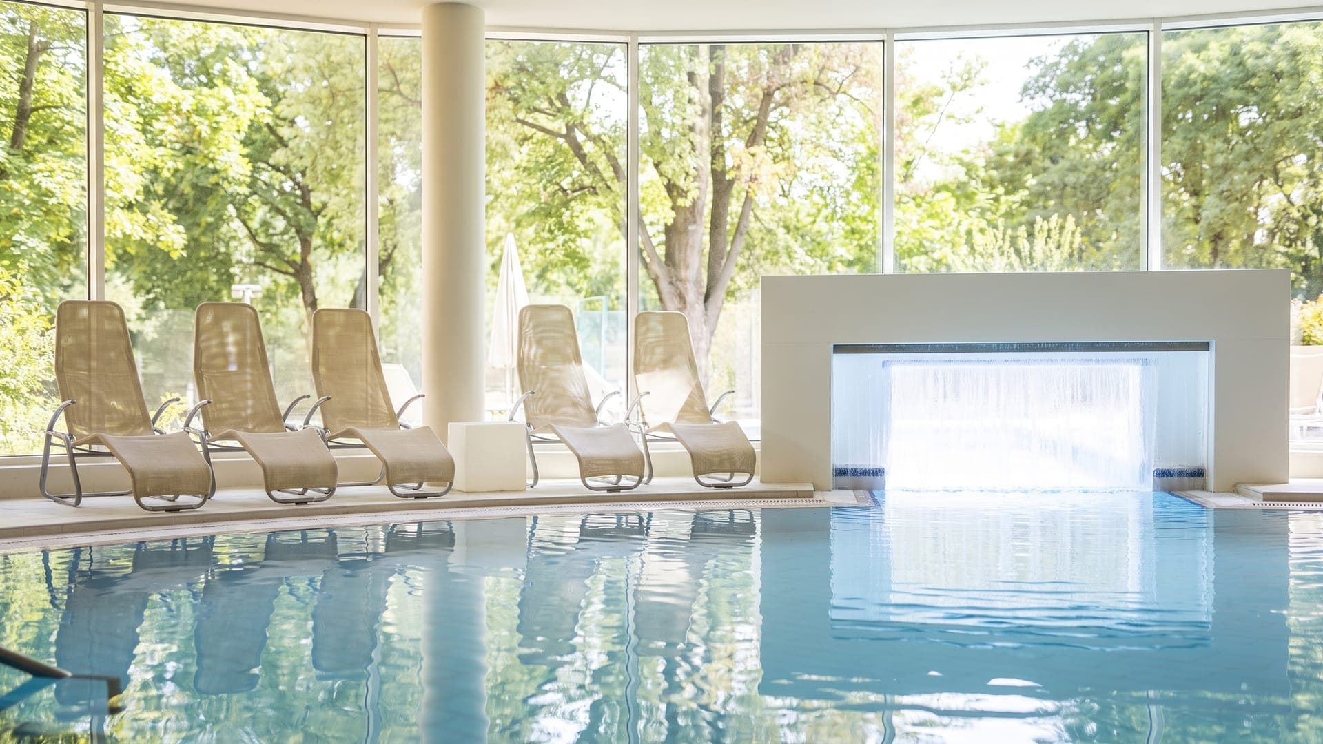 Indoor pool with a wide sheet of falling water, wicker loungers and a glass wall onto the trees
