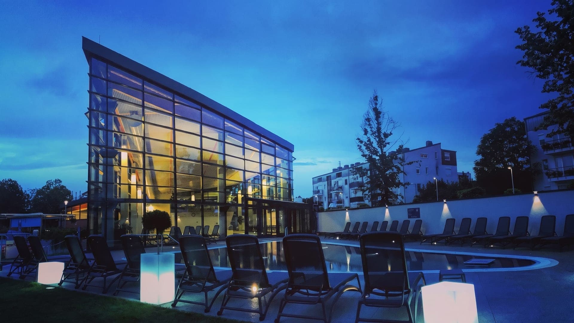 Glass-fronted bath building lit at dusk, loungers set out in front