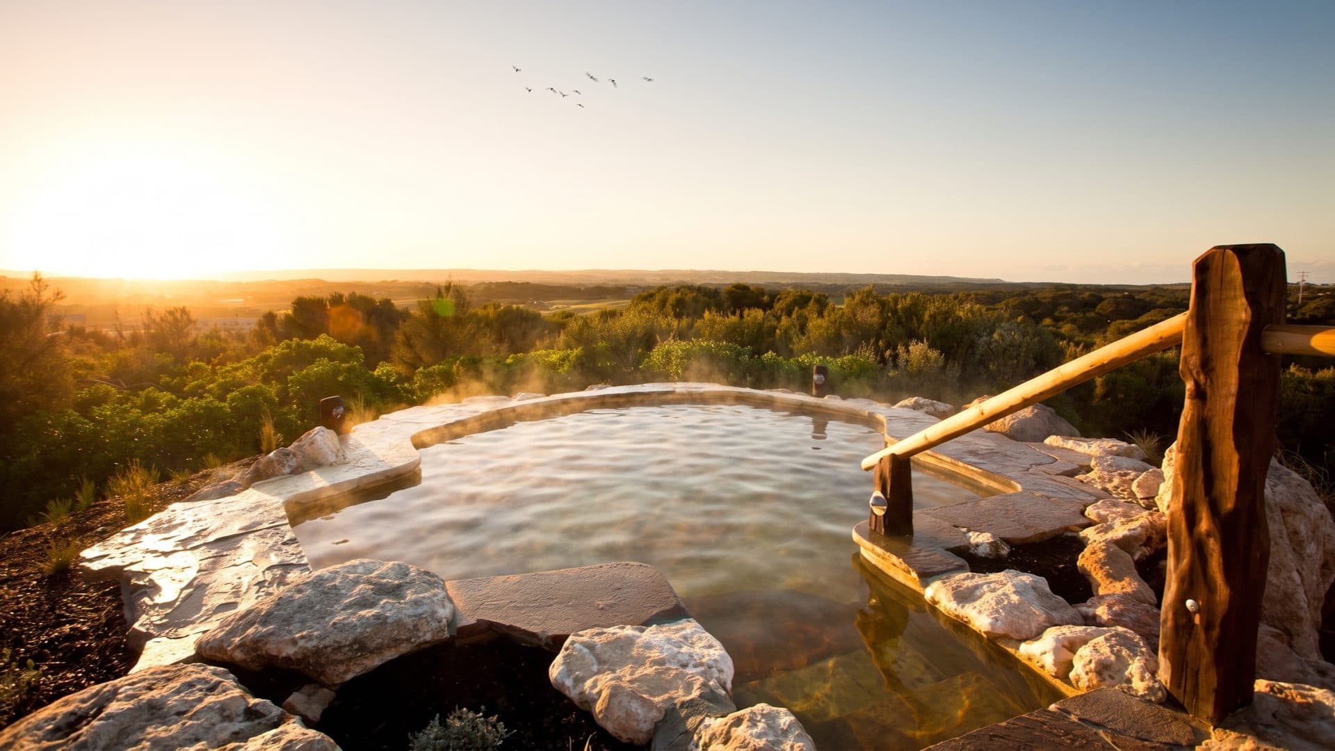Steaming circular hilltop bathing pool ringed with stone at sunset, looking out over coastal scrub and rolling country.