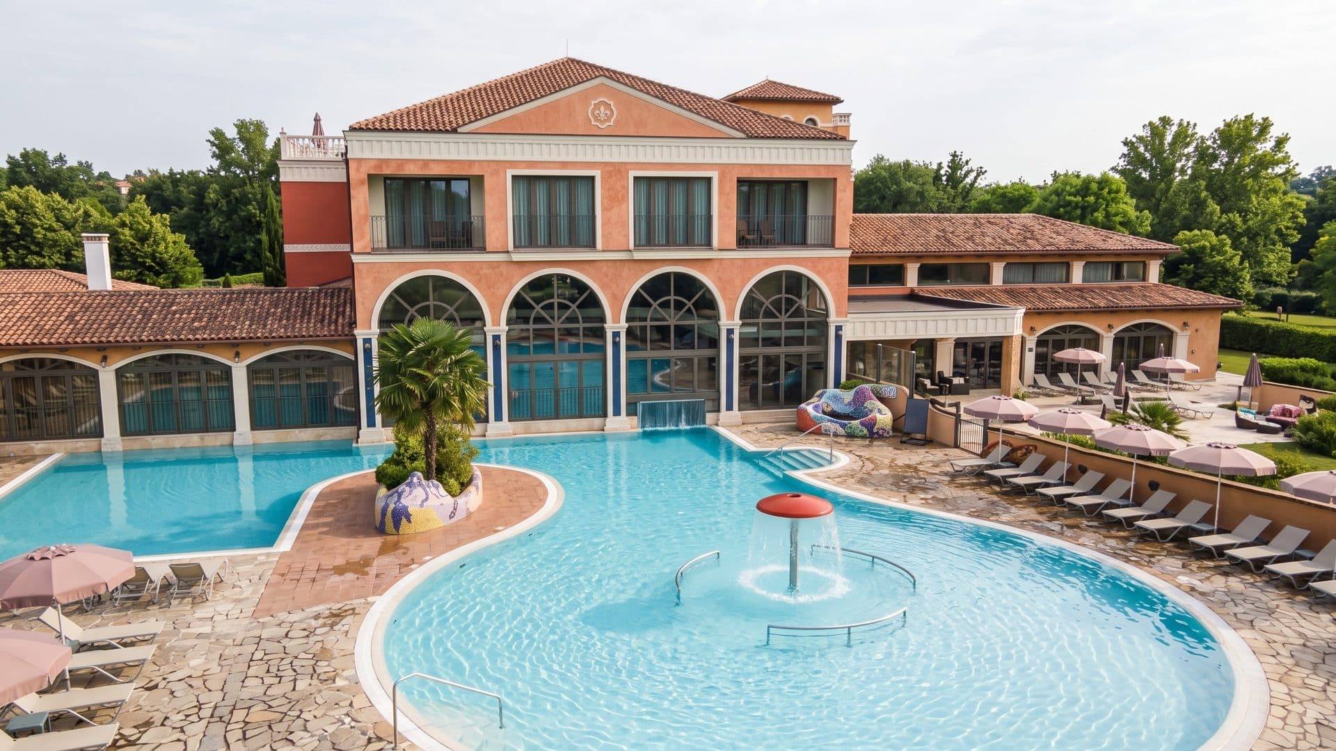 A curving outdoor pool with a mushroom fountain in front of the arched terracotta building of the Plaza Beach Resort in Presov