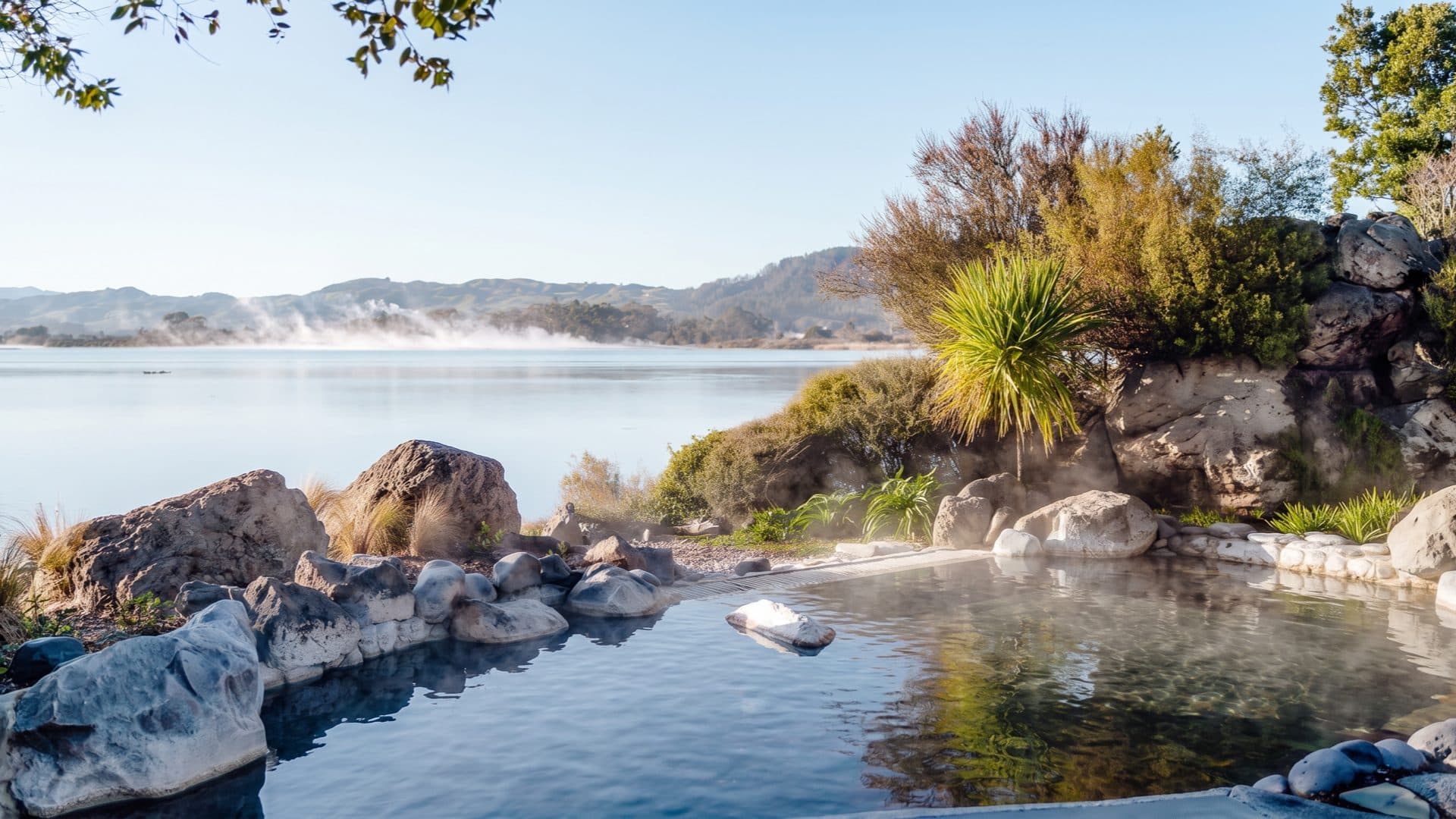 Boulder-edged thermal pool at the shore of a steaming lake, with native plants and low hills beyond in the morning light.