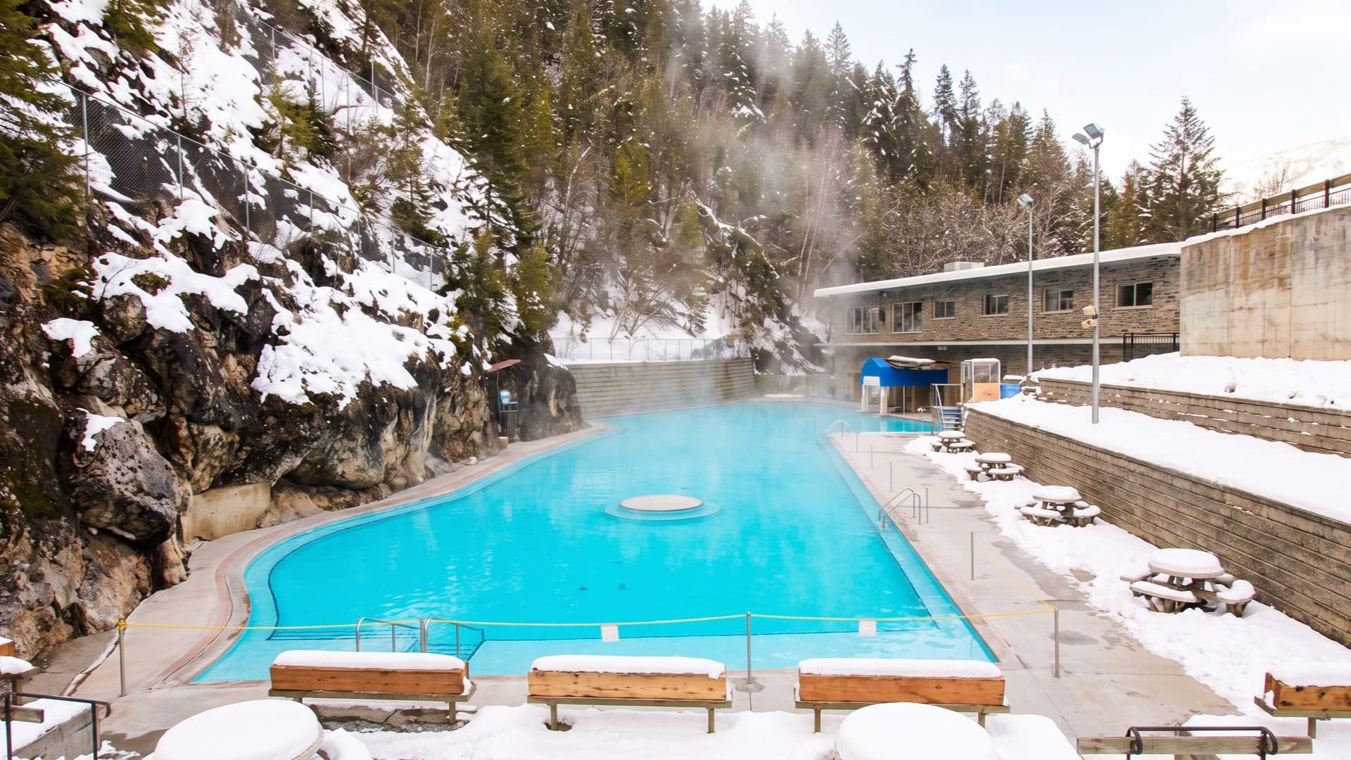 Long curving turquoise hot pool in a snowy rock canyon, steam rising against snow-laden pines and a low bathhouse.